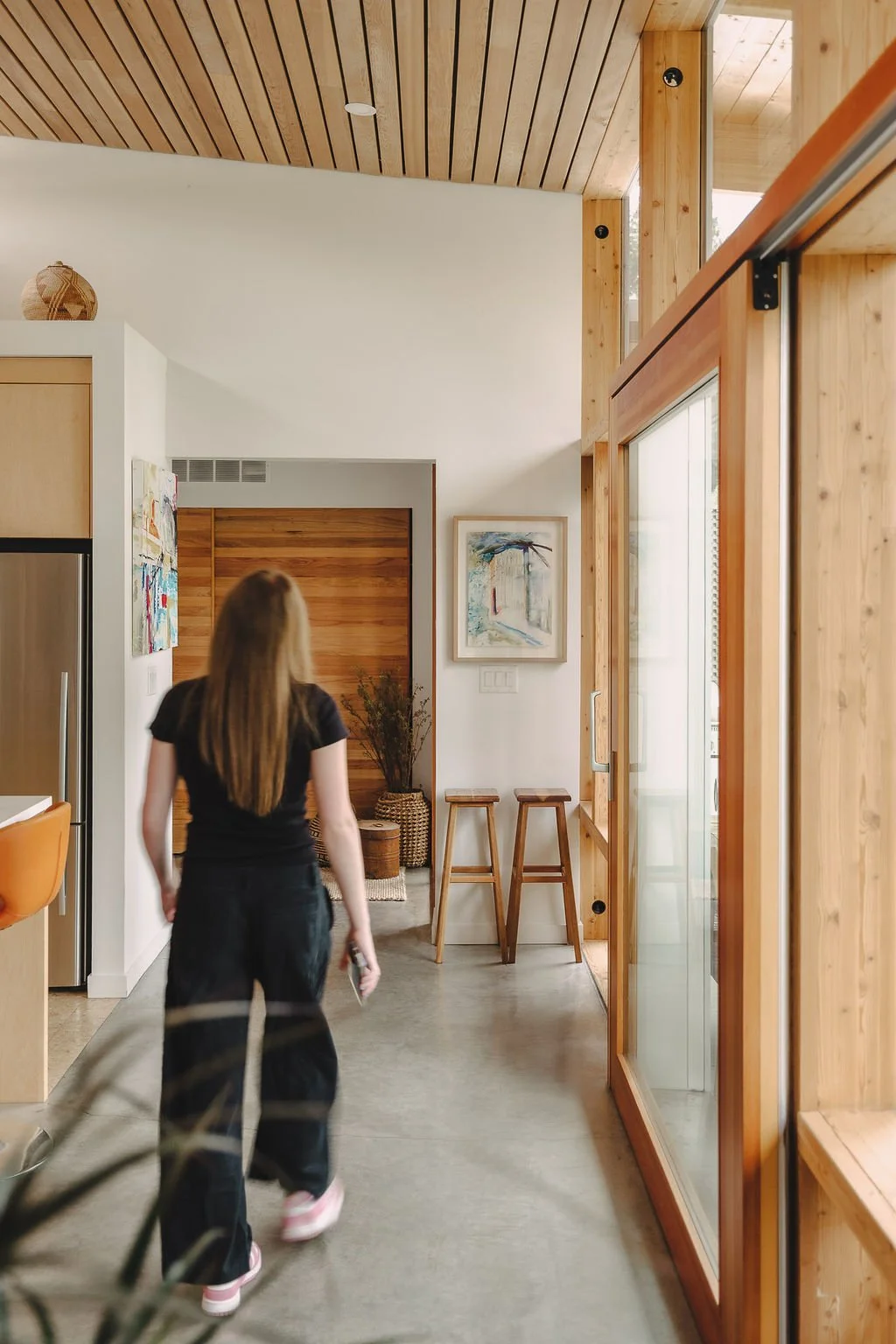 A woman walking inside a modern, wooden interior home, with art on the walls and natural light coming through large windows.