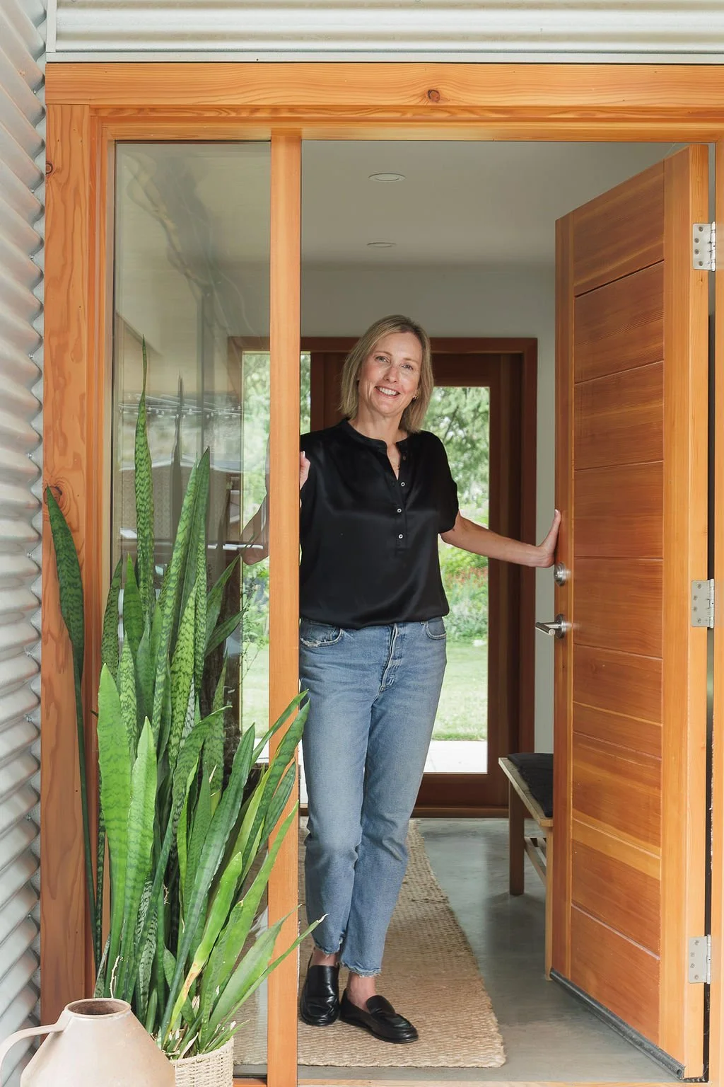 Woman standing in doorway, smiling, wearing black blouse, blue jeans, and black loafers, with a tall green plant and a ceramic vase nearby.