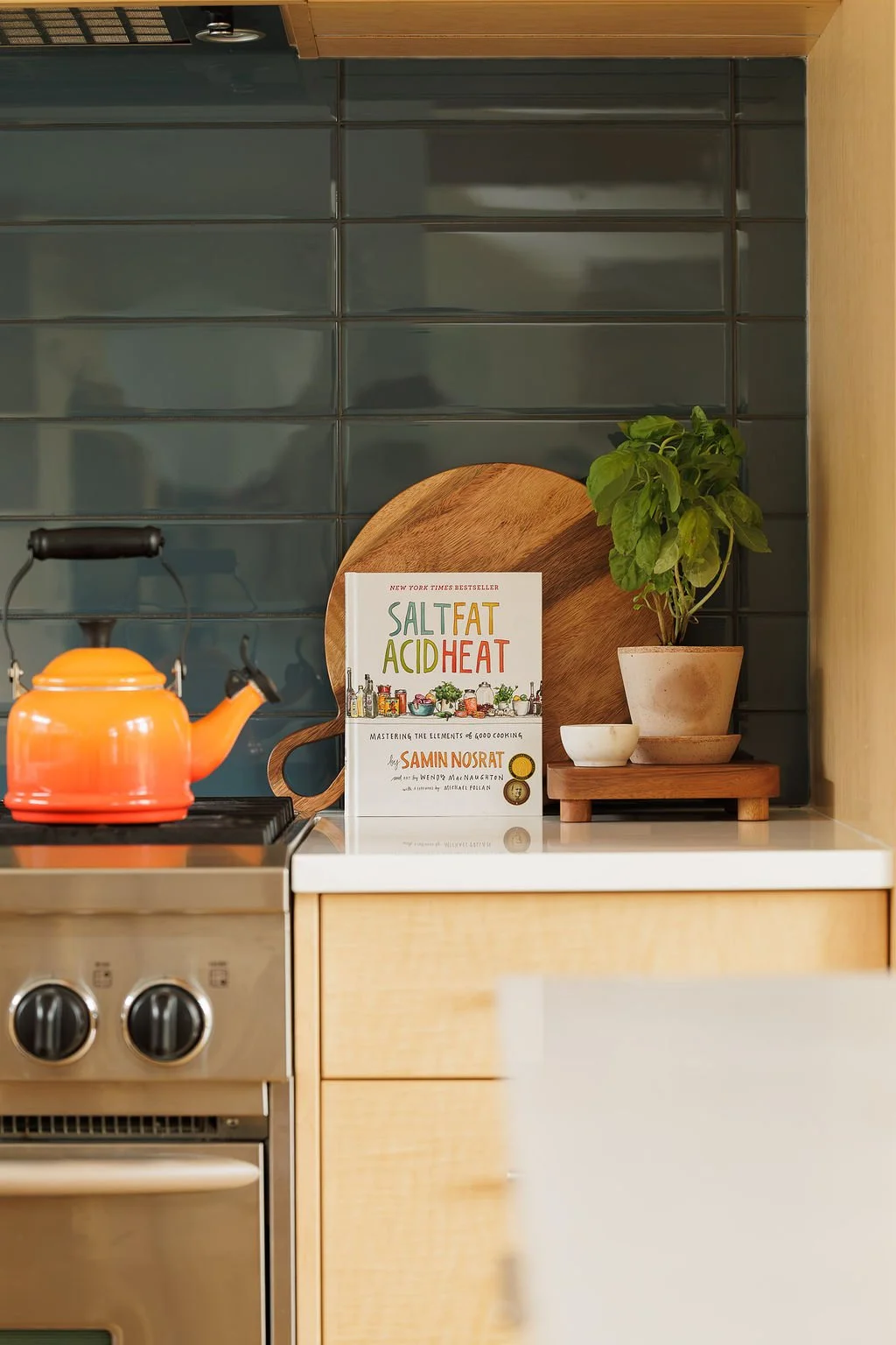 Kitchen countertop with a potted basil plant, a wooden cutting board, a white bowl, and a cookbook titled "Salt Fat Acid Heat" by Samin Nosrat. An orange kettle is on the stove in the foreground, and a dark tiled wall is in the background.