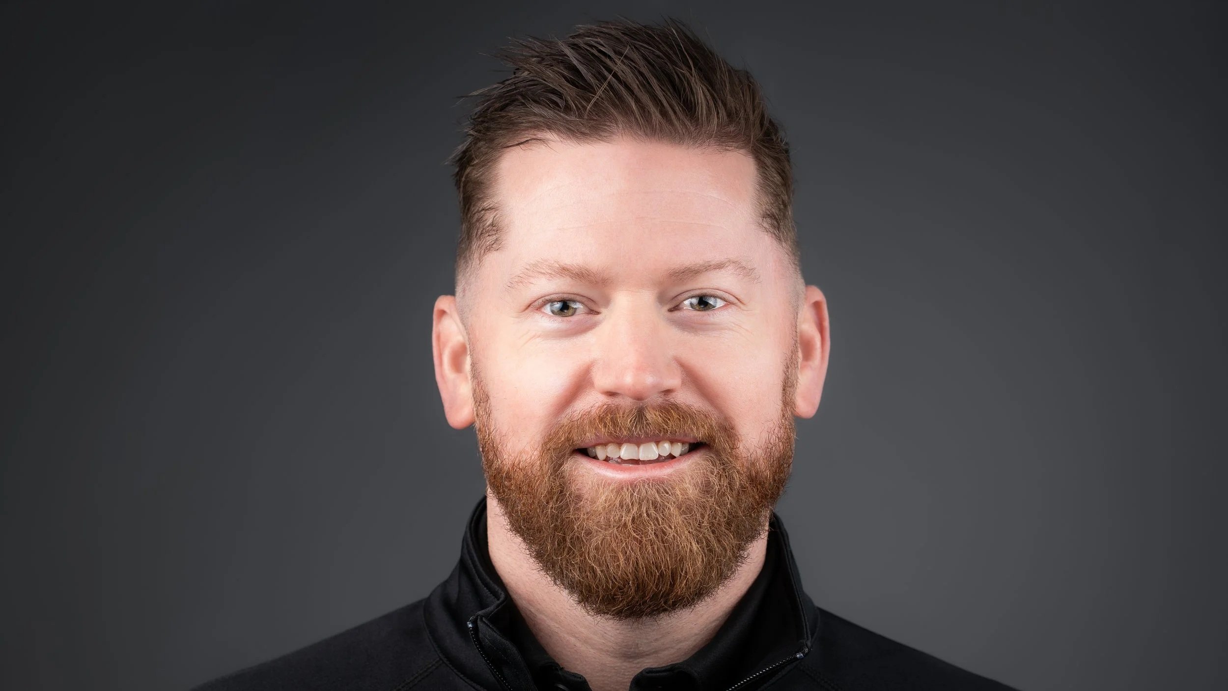 Headshot of a smiling man with red hair and beard, wearing a black zip-up jacket, against a dark gray background.