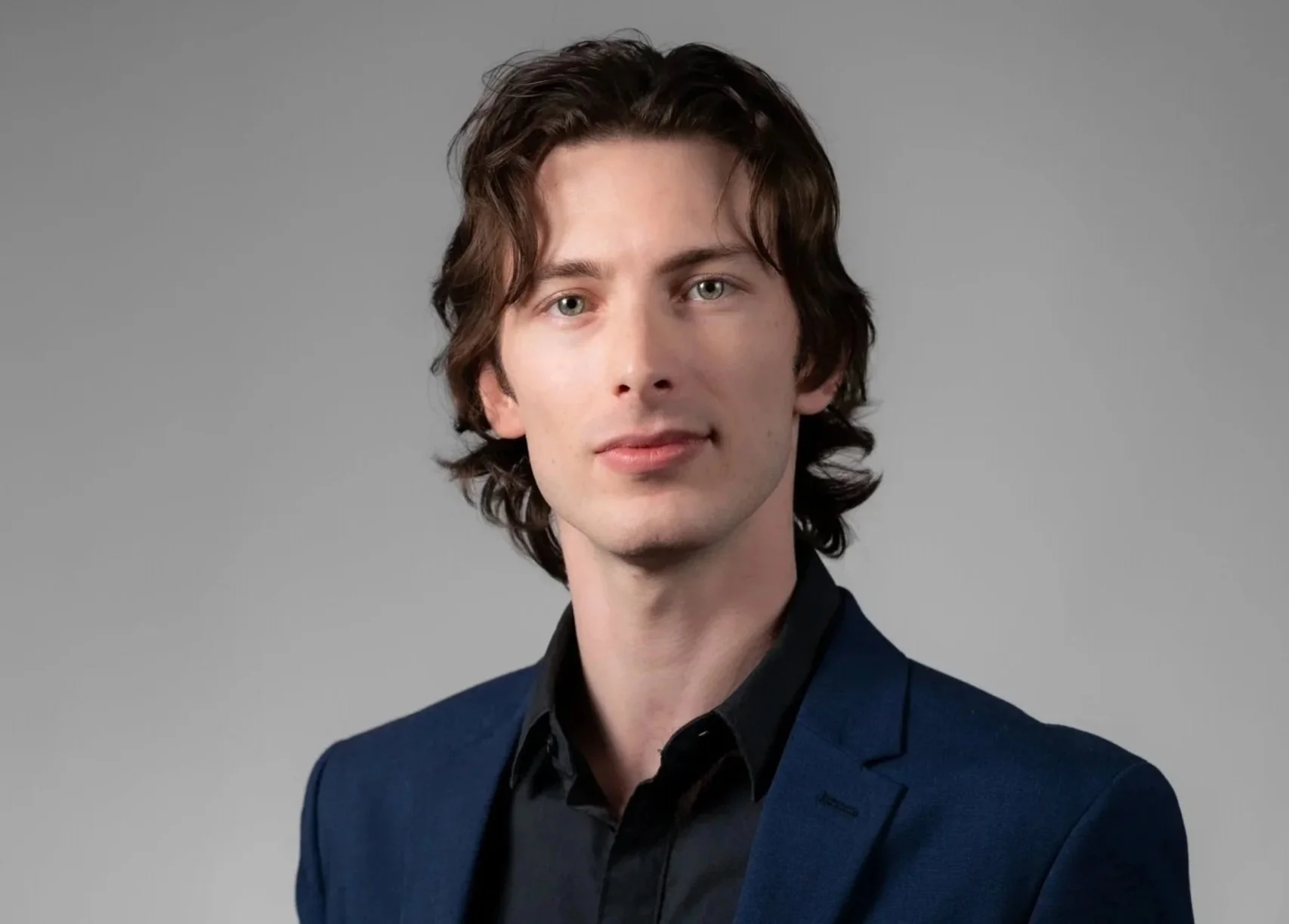 A man with wavy brown hair wearing a dark blue blazer and black shirt posed against a plain gray background.