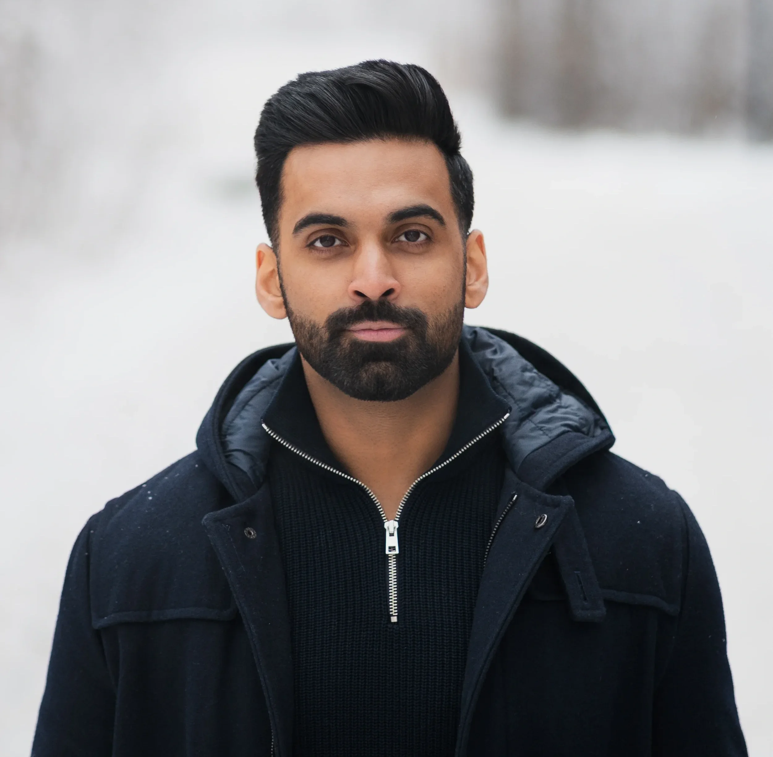 A man with dark hair and a beard standing outdoors in winter, wearing a dark jacket and a zip-up sweater, with a snowy background.