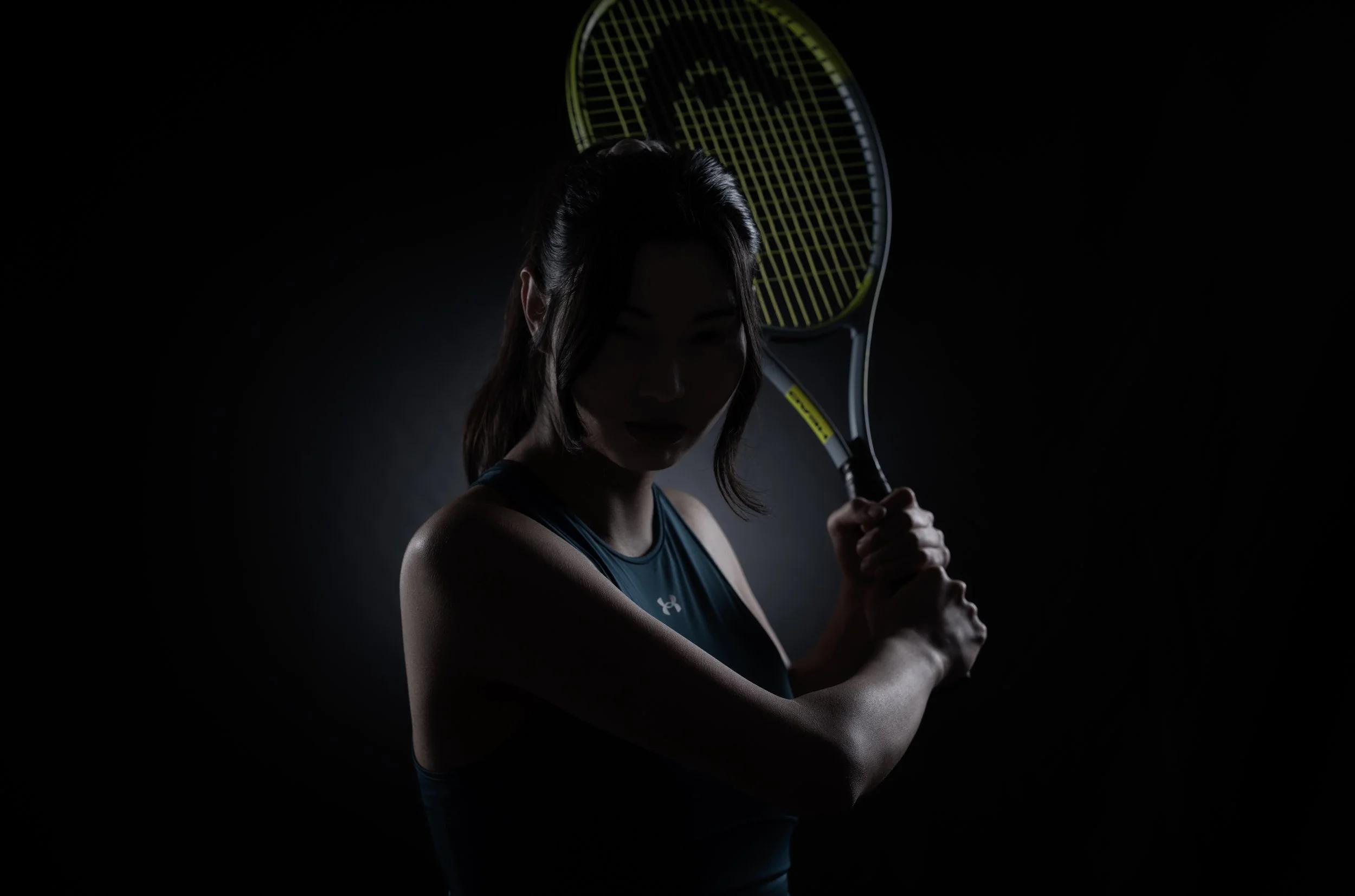 A woman in athletic wear holding a tennis racket with a dark background and subtle lighting highlighting her silhouette.