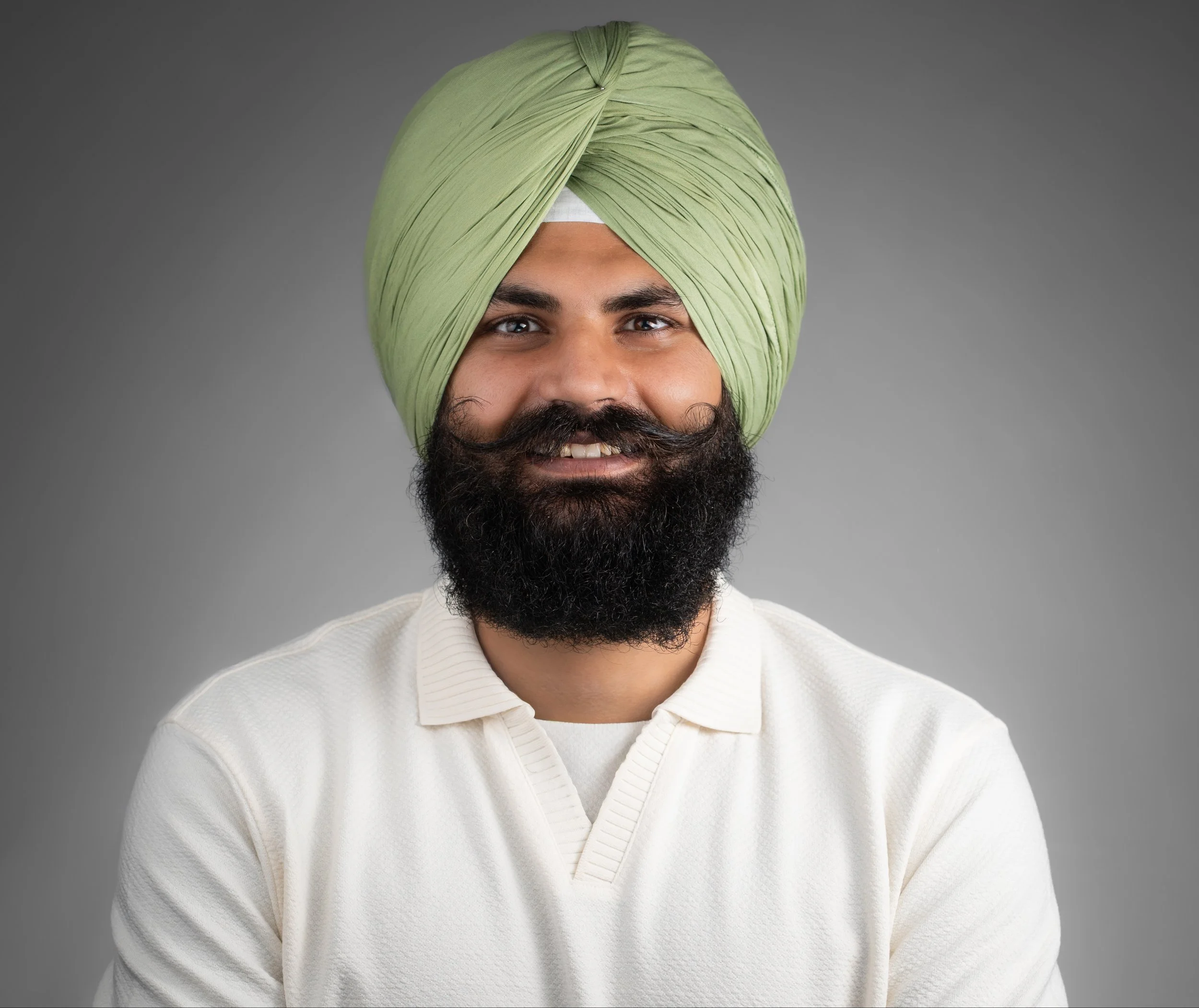 Portrait of a man with a green turban, a full beard, and a mustache, smiling against a gray background.