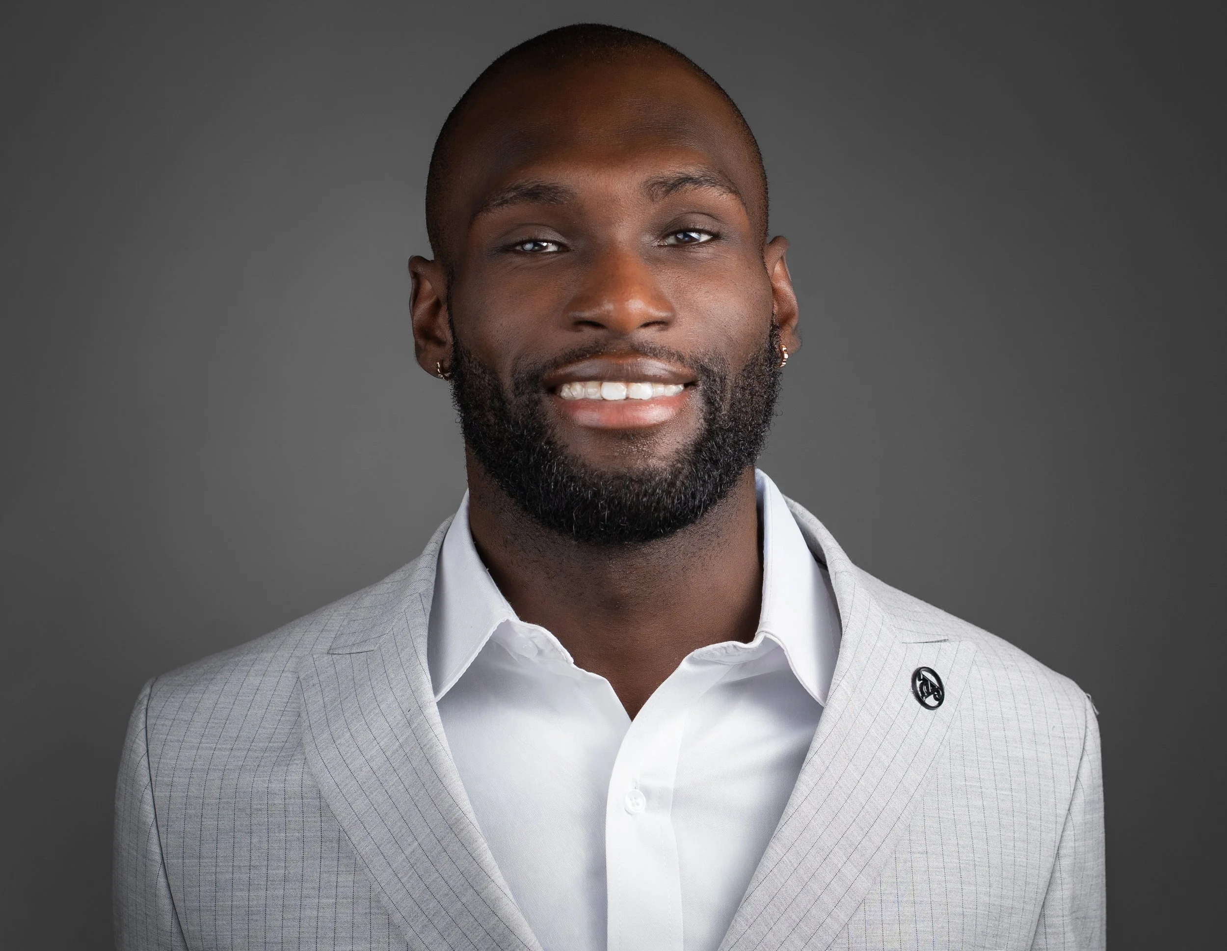 A smiling man with a beard and earrings wearing a light gray suit and white shirt against a dark gray background.