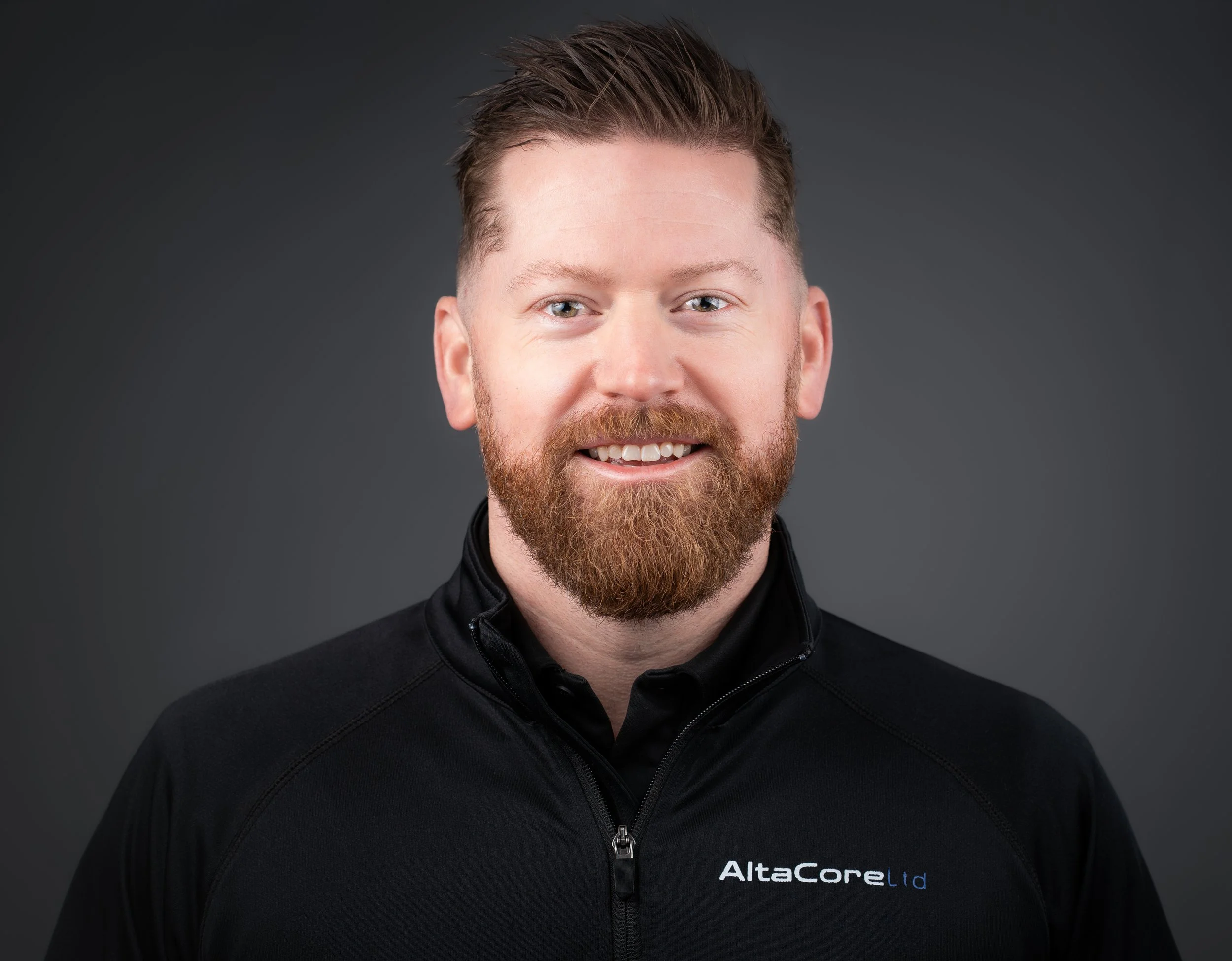Headshot of a man with red hair and beard, smiling, wearing a black jacket with a company logo 'AltaCore Ltd' on it, against a dark gray background.