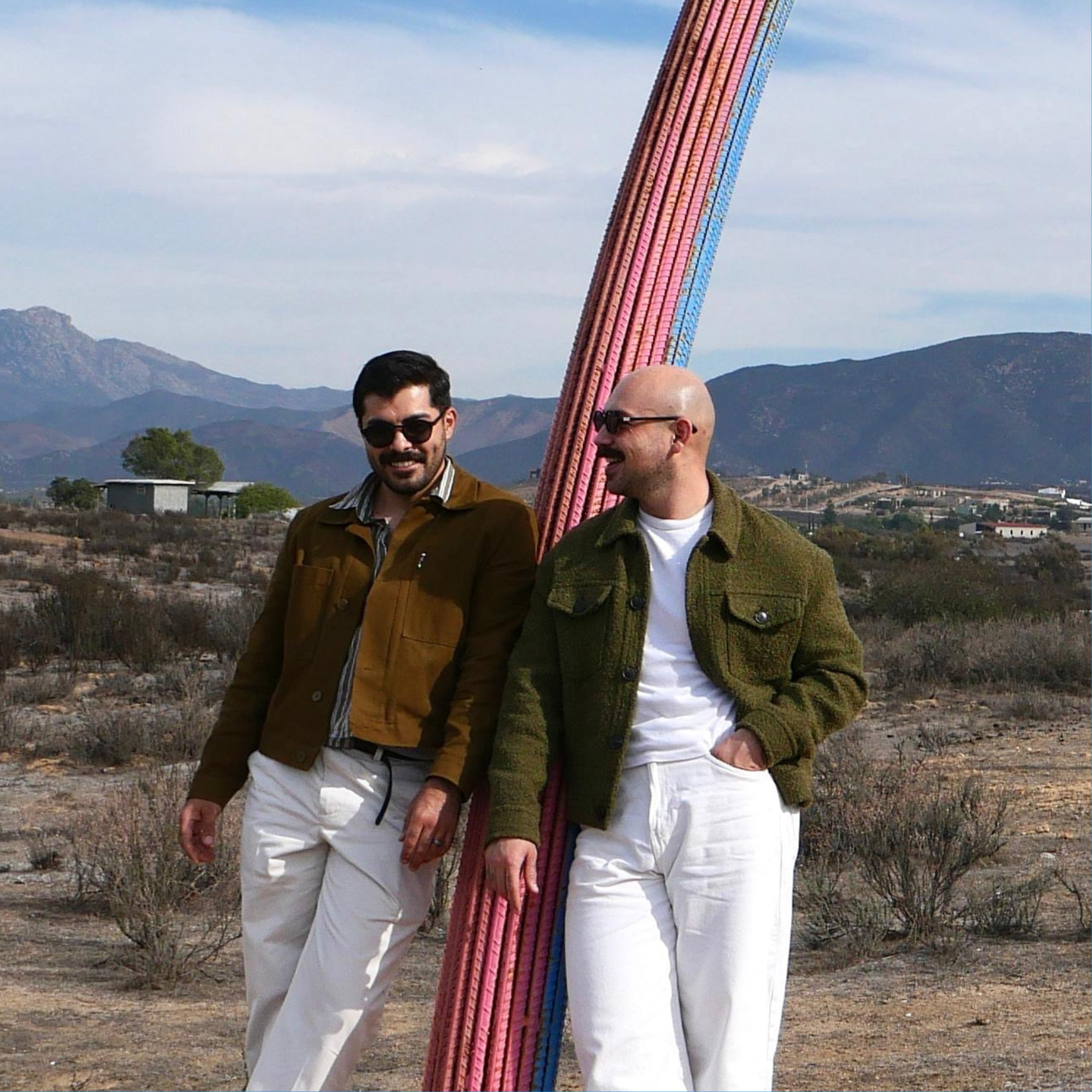 Dos hombres sonrientes con gafas de sol de pie en un paisaje desértico, junto a una escultura de arco colorida, con montañas y un cielo parcialmente nublado al fondo.
