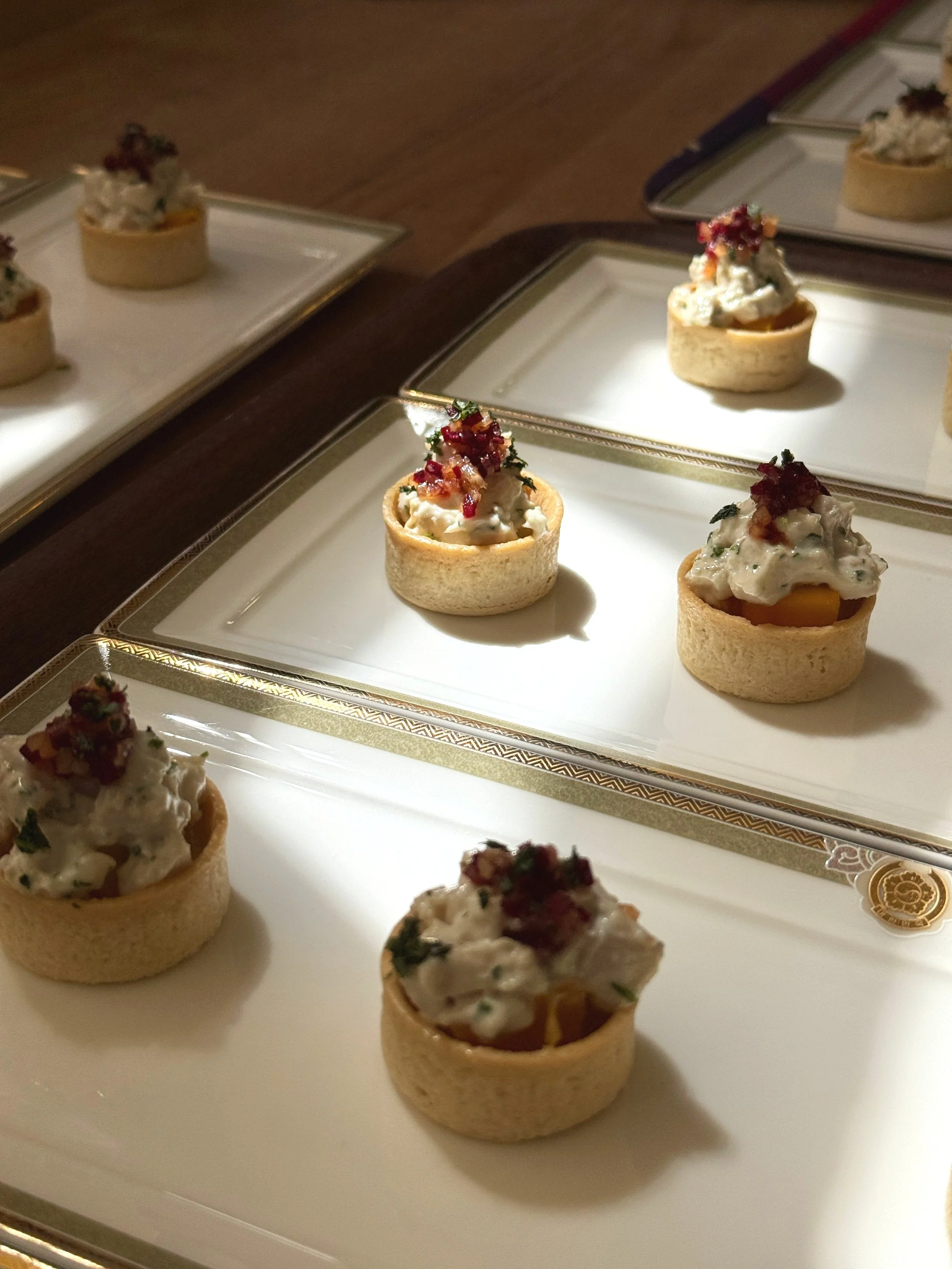 Appetizer tartlets with a cream topping and garnished with herbs and red bits, arranged on decorative white and gold trays.