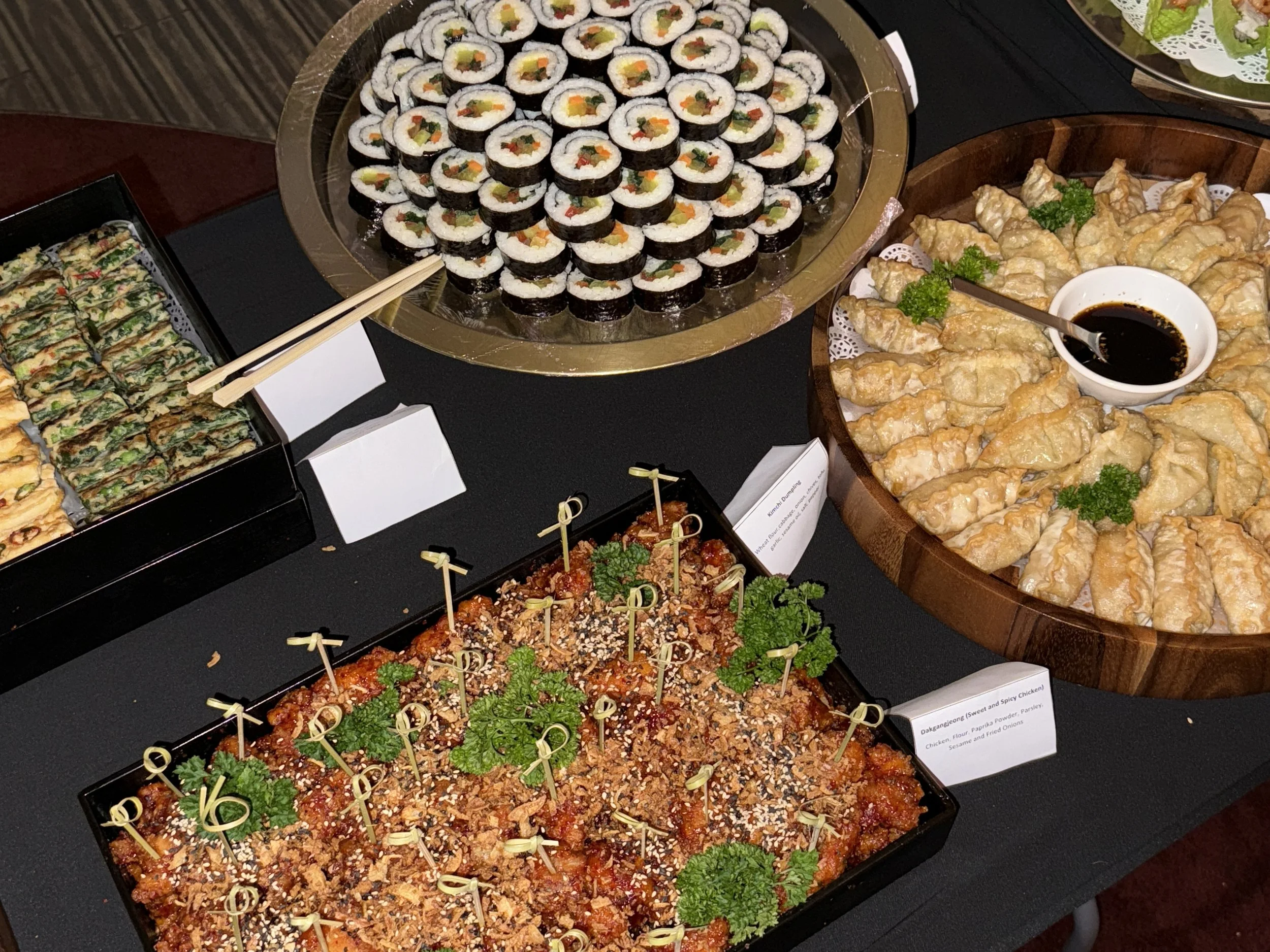 A variety of Asian dishes on a buffet table, including kimbab, fried dumplings with dipping sauce, and a tray of spicy chicken bites garnished with parsley and sesame seeds.