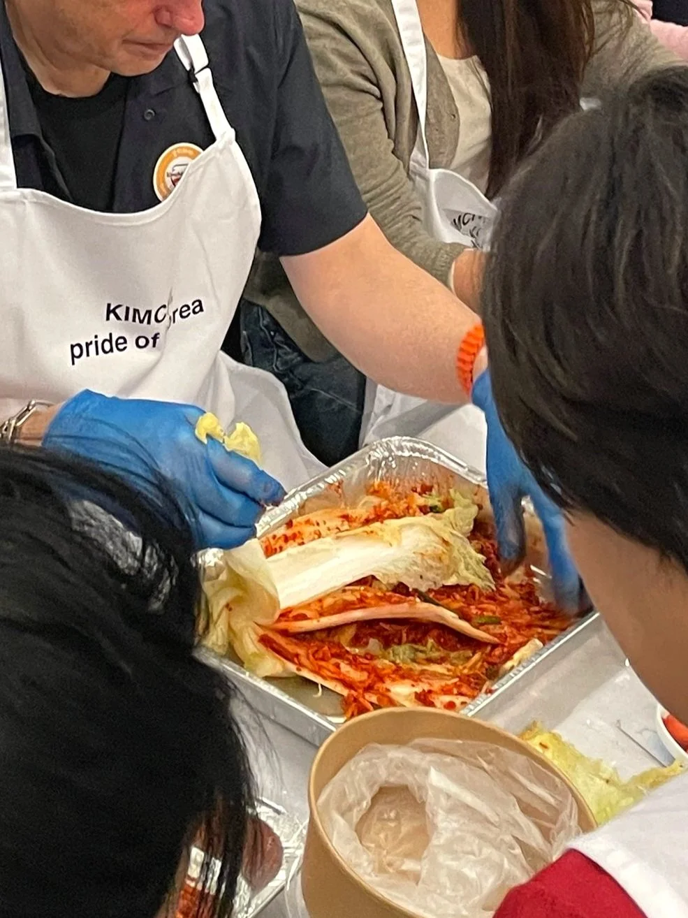 People gathered around a tray of kimchi, preparing or serving it. Some individuals are wearing aprons and gloves, suggesting a communal meal or food preparation event.