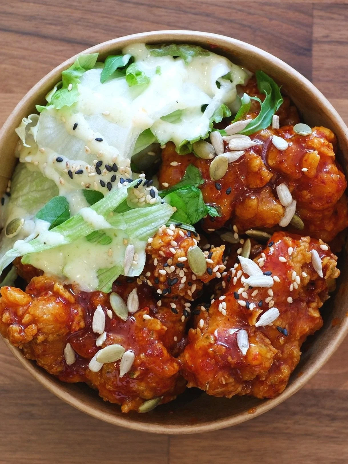 Bowl of honey garlic chicken bites topped with sunflower seeds, black sesame seeds, and chopped lettuce, served on a wooden surface.