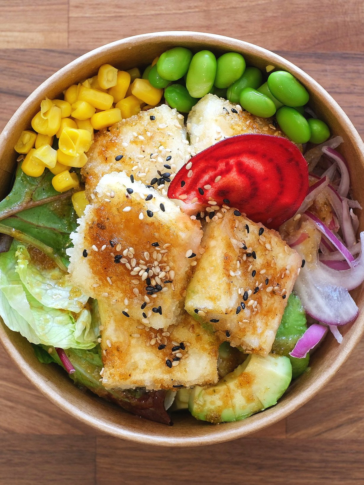 A salad bowl containing fried fish pieces topped with sesame seeds, mixed greens, avocado slices, sliced tomato, corn, edamame, and red onion.