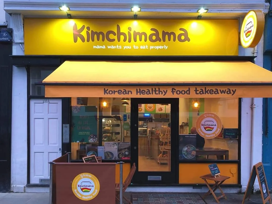 Korean takeaway restaurant named Kimchimama with yellow signage and awning, storefront displaying welcoming interior, and signs indicating Korean healthy food takeout.