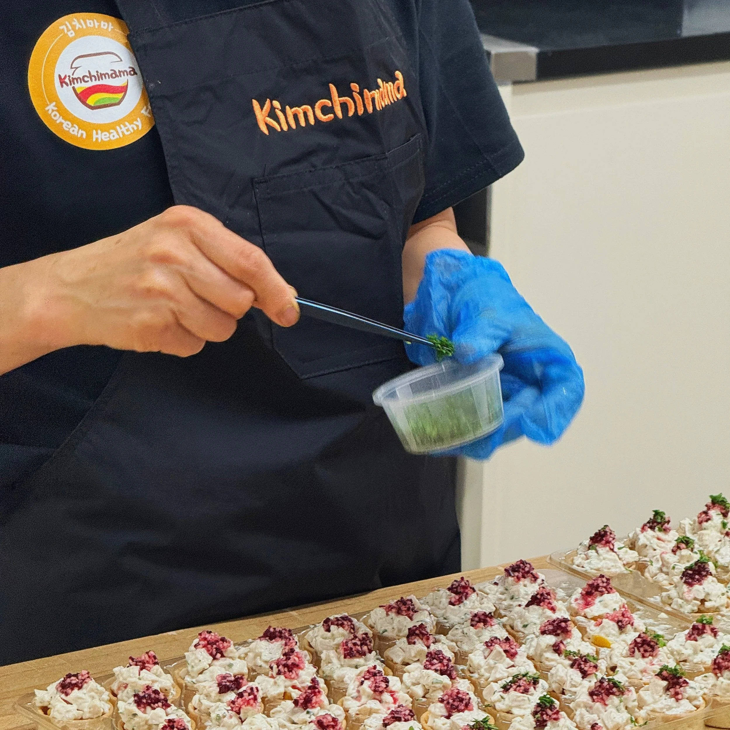 A person wearing a dark apron labeled 'Kimchimama' and a patch that says 'Korean Healthy' is decorating small bite-sized food items with green herb using tweezers in a plastic container, with more decorated food items on a wooden surface in front.