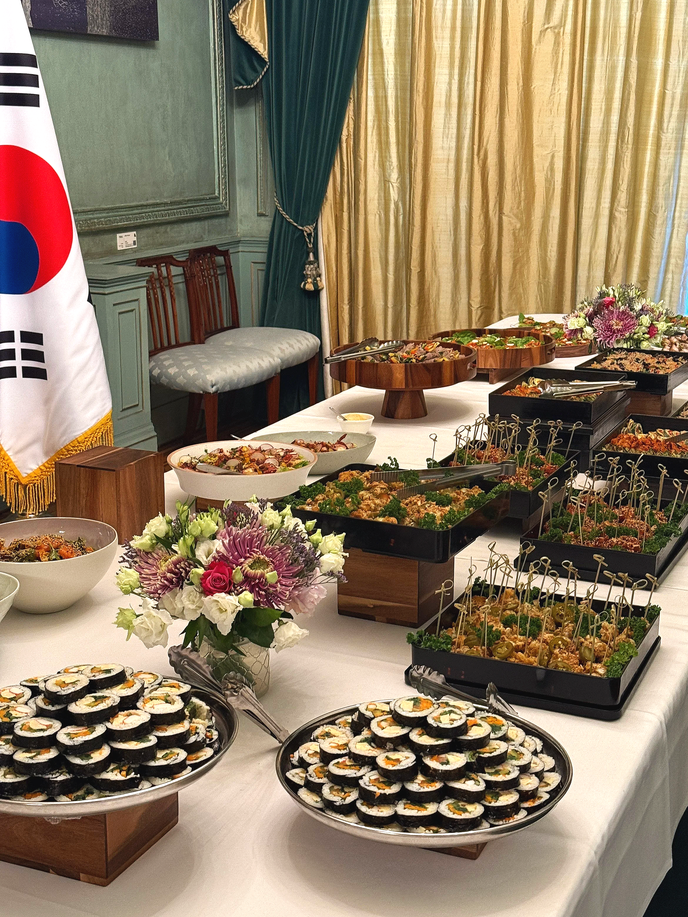 A catering buffet table with kimbab, salads, and appetizer trays, decorated with flowers in a formal setting with gold curtains and green drapes.