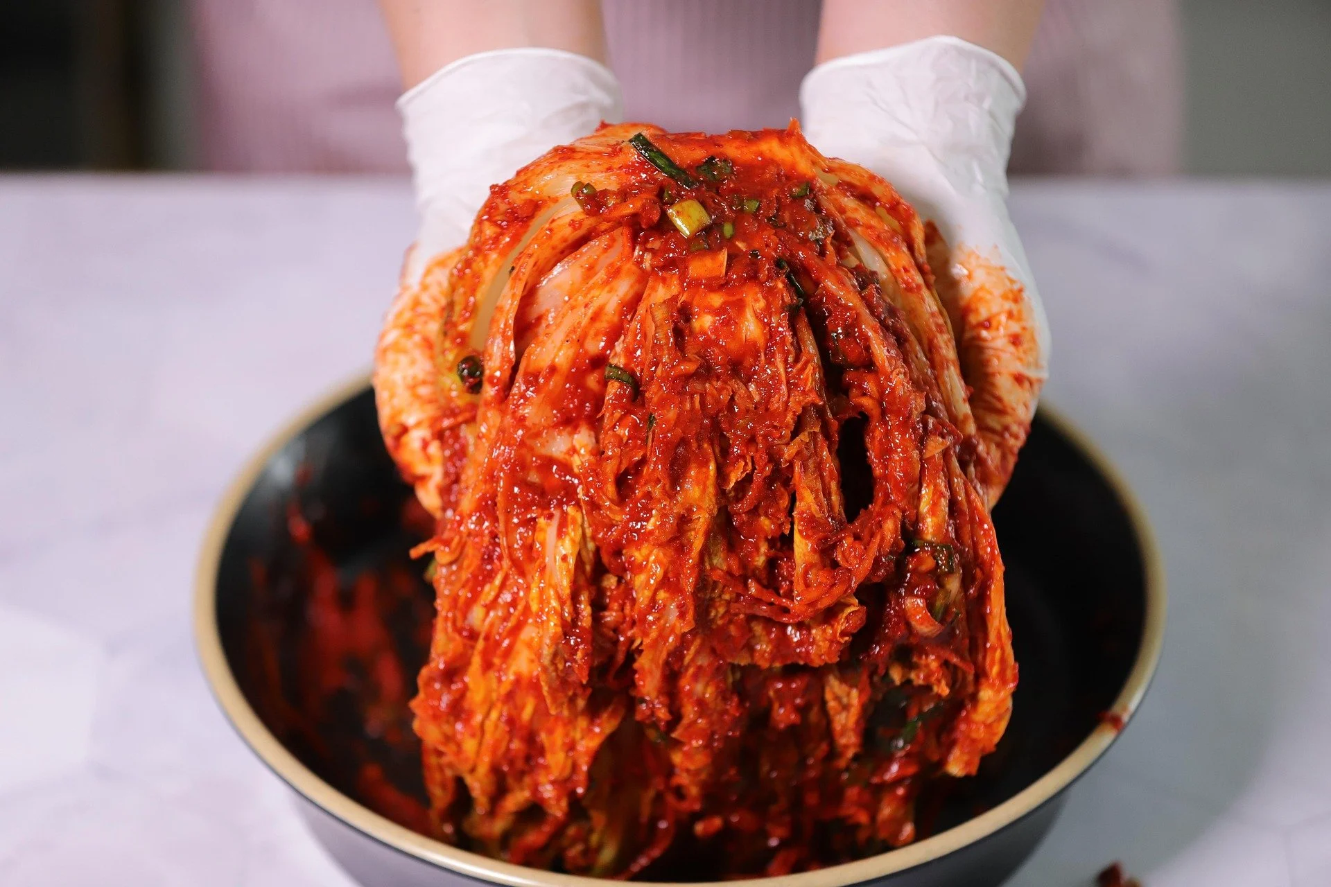 Person wearing white gloves holds a large bowl of kimchi, a fermented Korean side dish made of cabbage with red pepper seasoning.