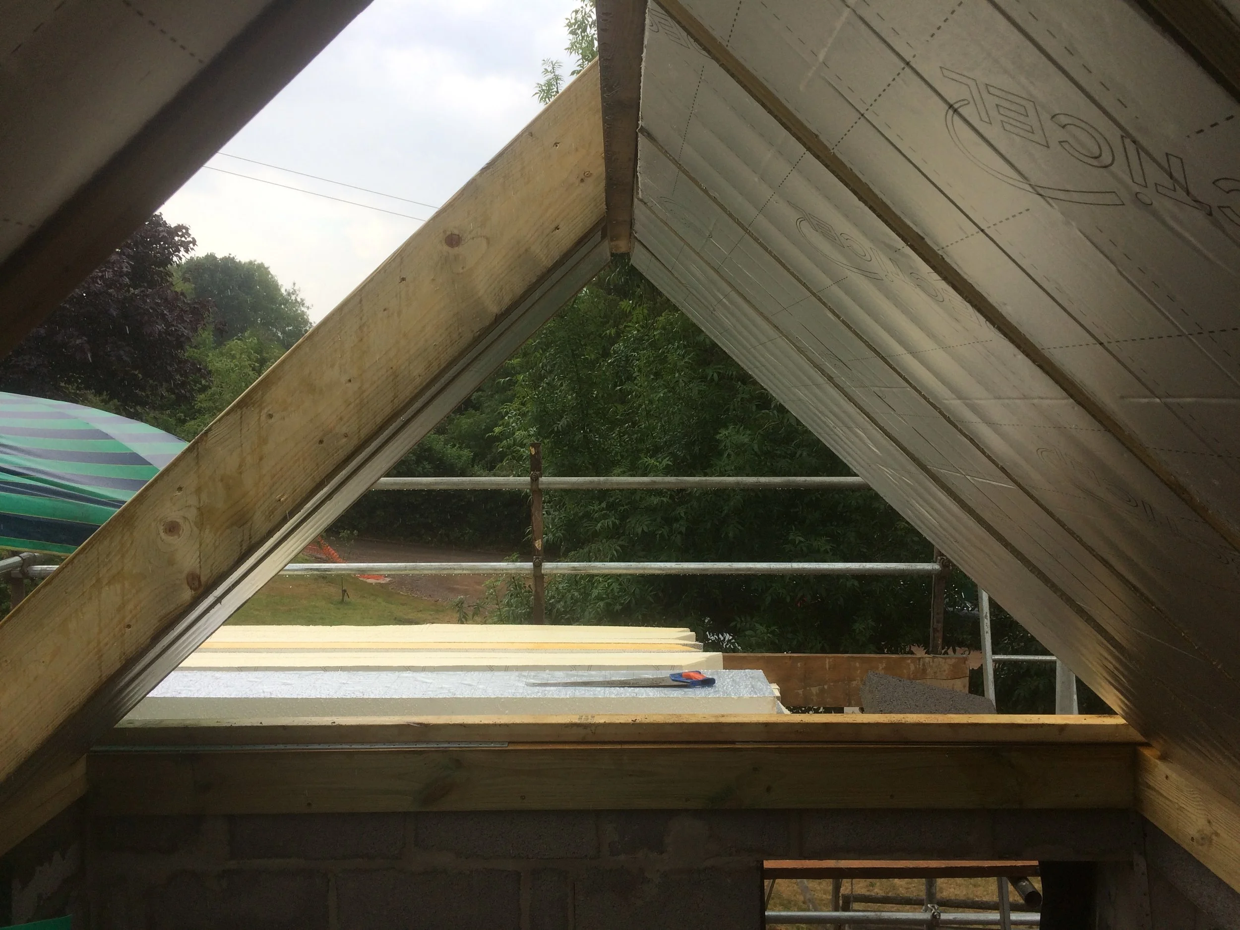 Construction site with roof framing and insulation being installed, view through an opening showing cloudy sky and green trees outside.