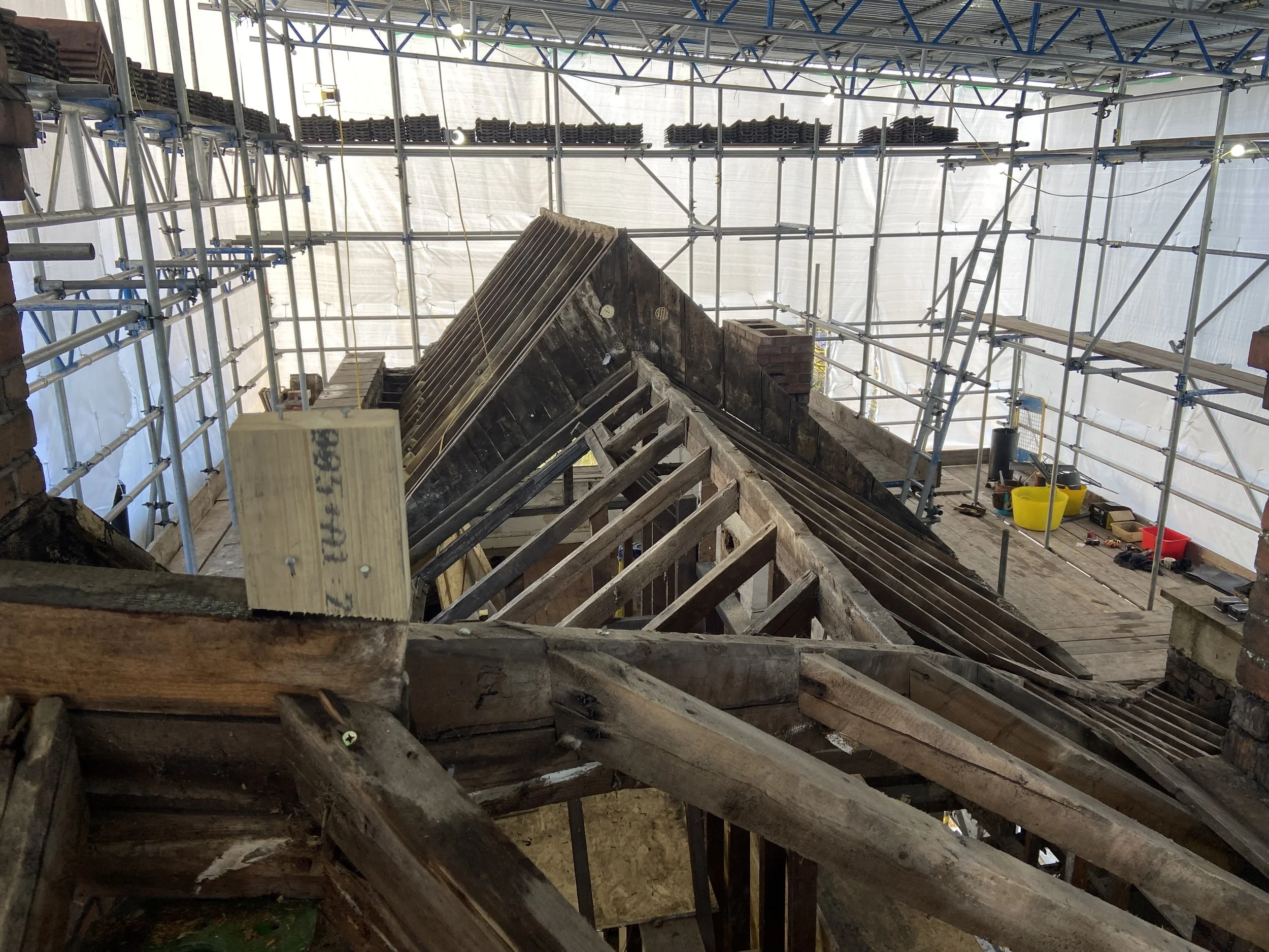 View of a house roof under renovation, with scaffolding and construction tools present, showing wooden framework and parts of roof structure.
