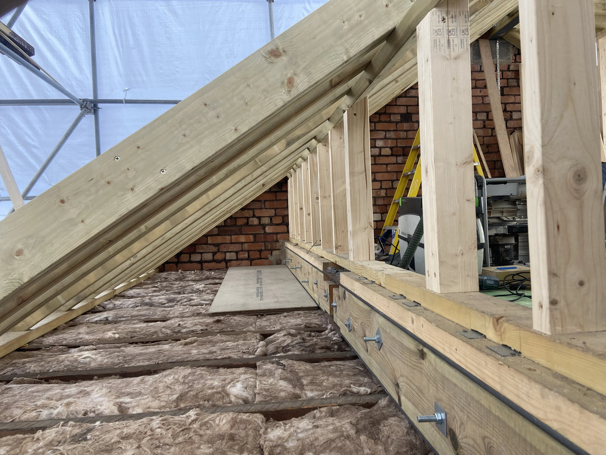 Construction site showing wooden framing and insulation for a building's roof, with a brick wall in the background and construction tools present.