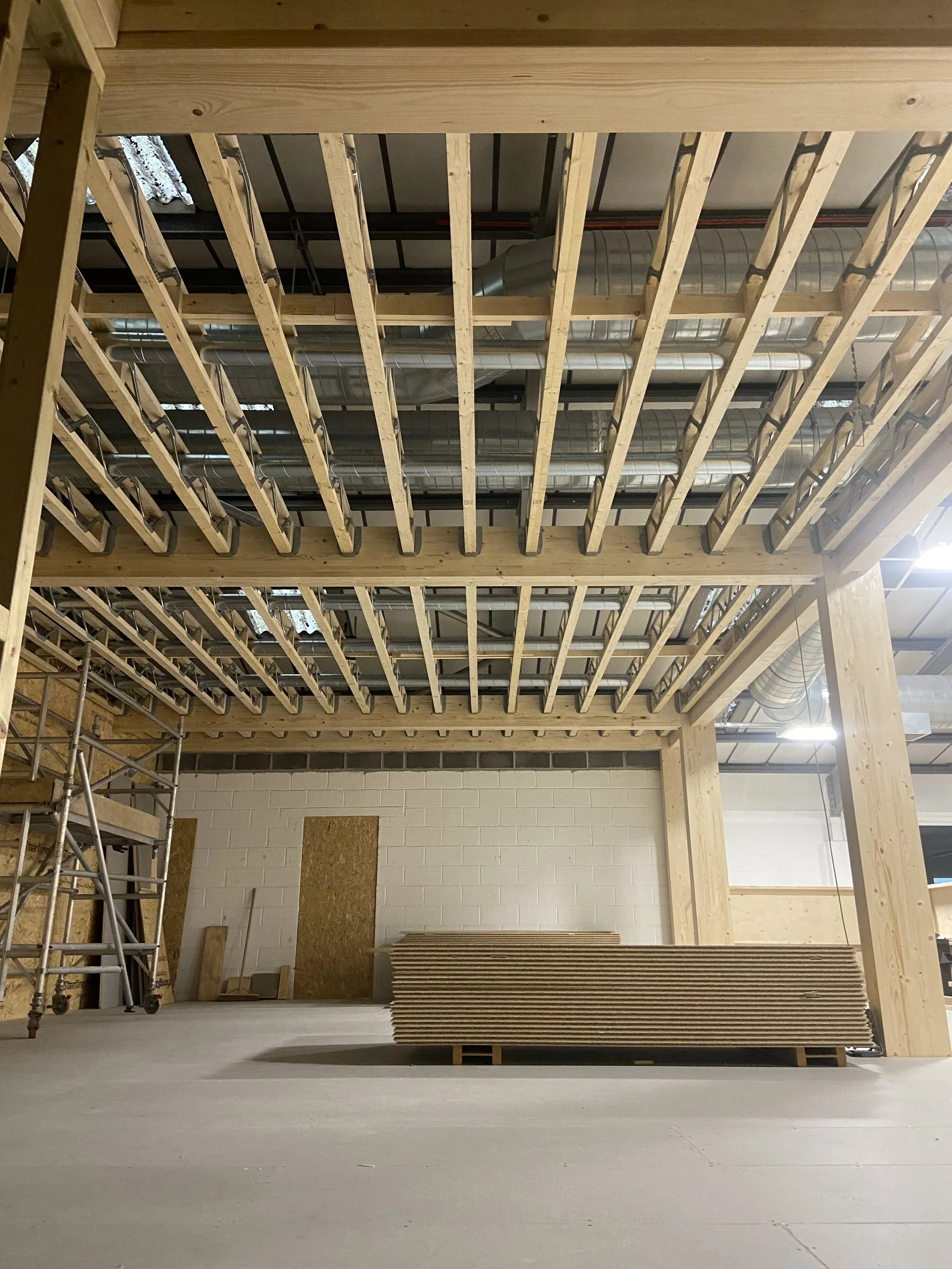 Construction site with exposed wooden framing for a ceiling or loft area and scaffolding on the left side, with some stacked plywood sheets on the floor, unfinished interior space.