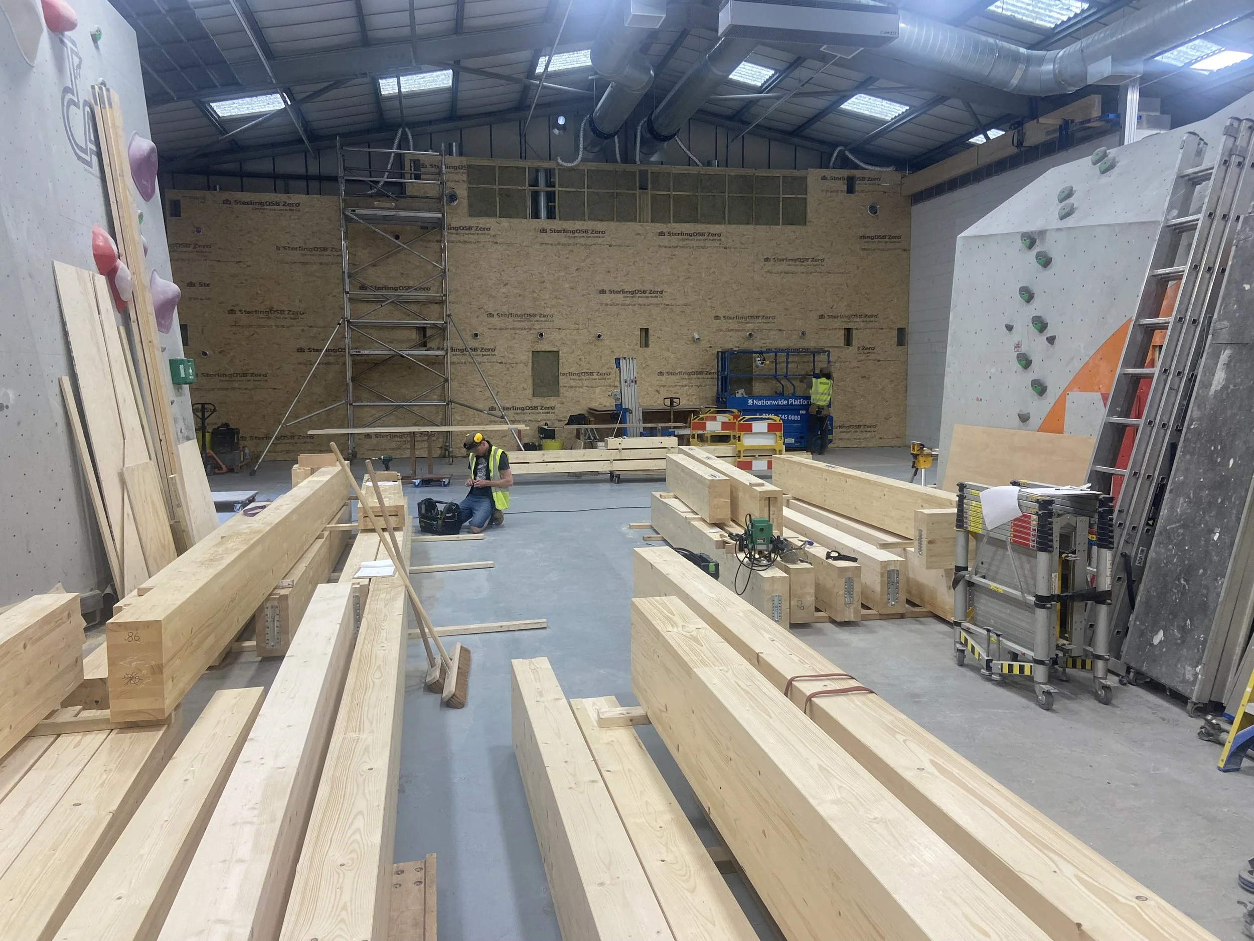Construction site of an indoor climbing gym with large wooden beams, scaffolding, and a climbing wall in the background, along with workers and construction equipment.