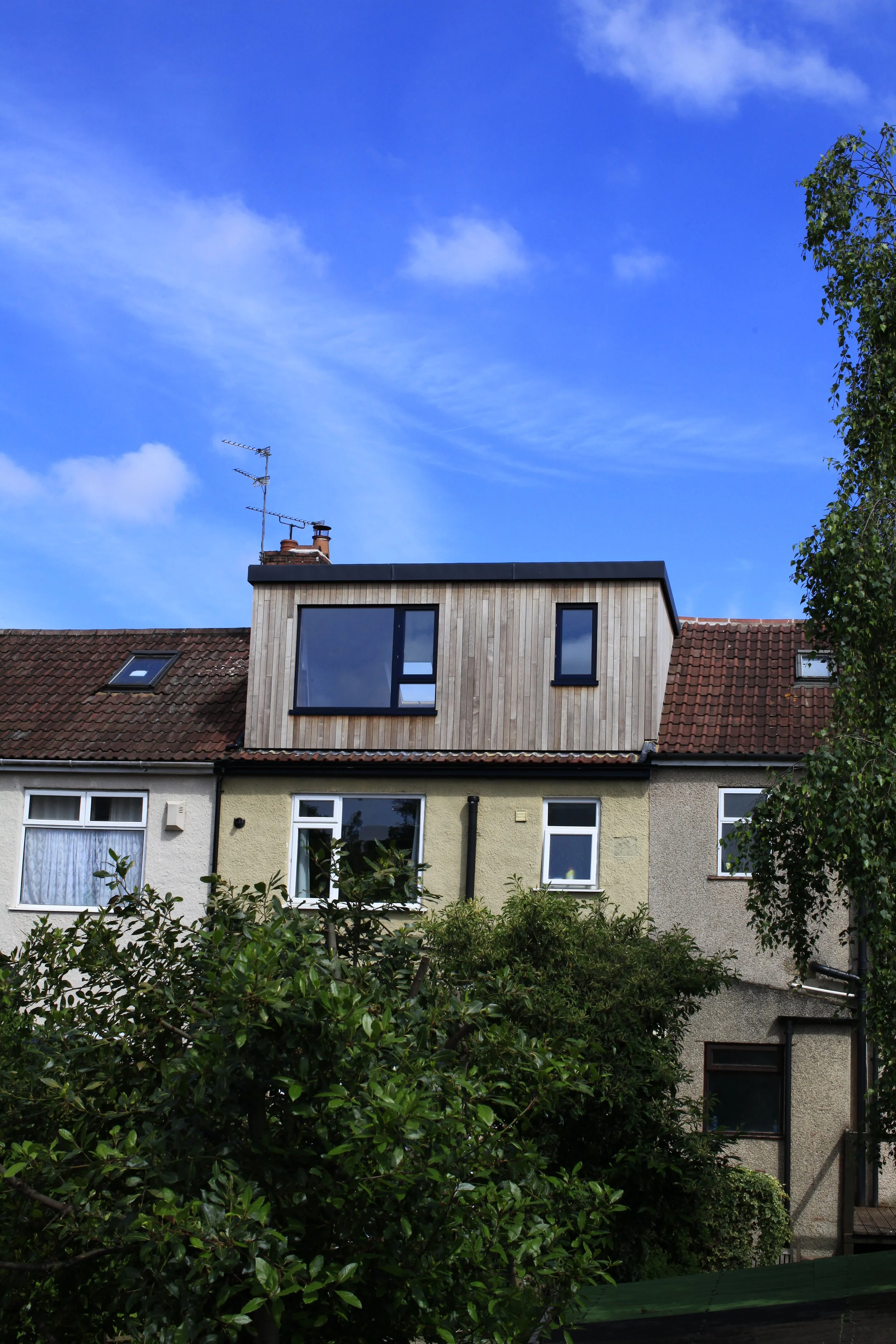 Apartment building with a new wooden dormer window extension on the roof, surrounded by green shrubbery and trees, under a blue sky with scattered clouds.