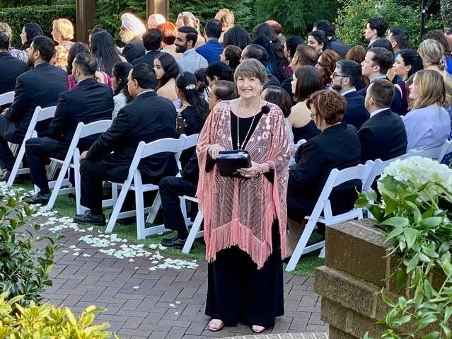 Officiant standing outdoors at a formal wedding with many guests seated behind her. She is smiling and holding a ceremony book. The event is a wedding with white chairs and flower petals.
