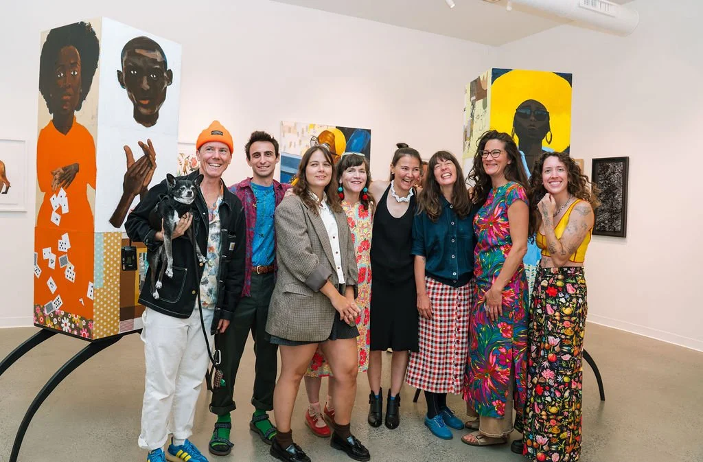A group of nine people, mostly women, smiling and posing together at an art gallery. One man is holding a small dog. The background features colorful modern artwork, including paintings of people with varying styles.