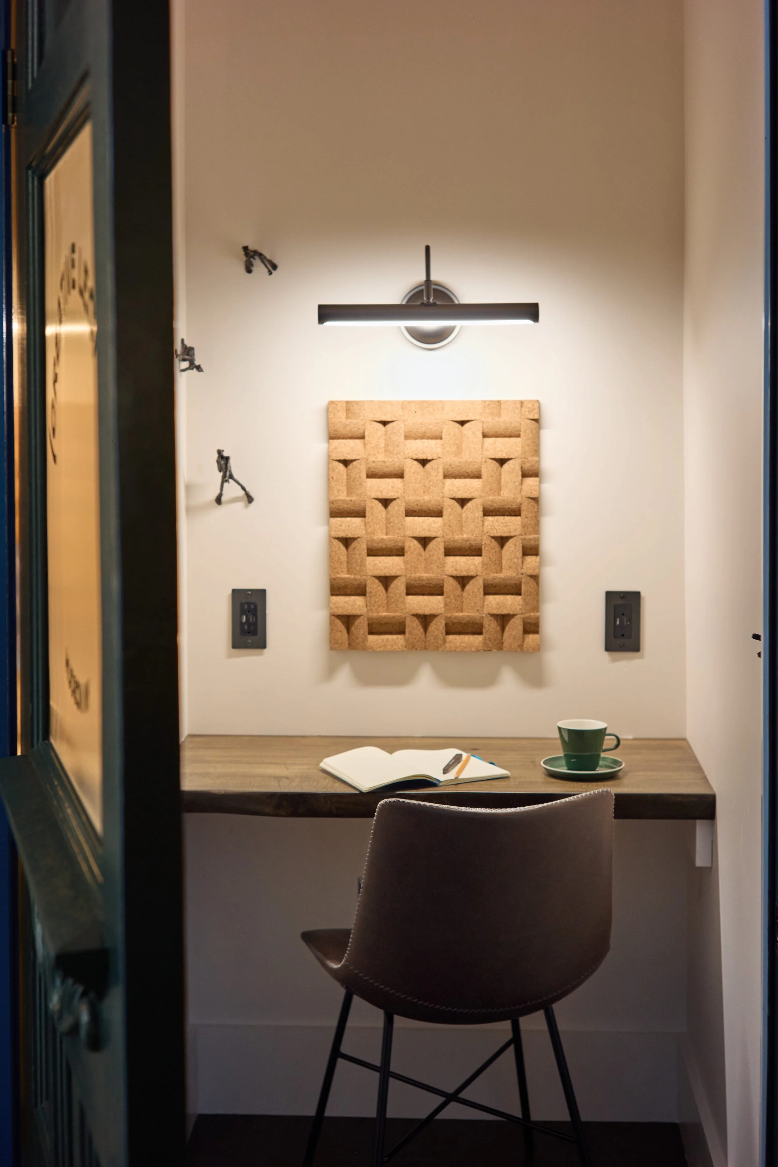A small, cozy workspace with a wooden desk, a gray chair, an open notebook, a green coffee cup and saucer, wall-mounted electrical outlets, and acoustic panels on the wall, illuminated by a black modern wall-mounted light fixture.