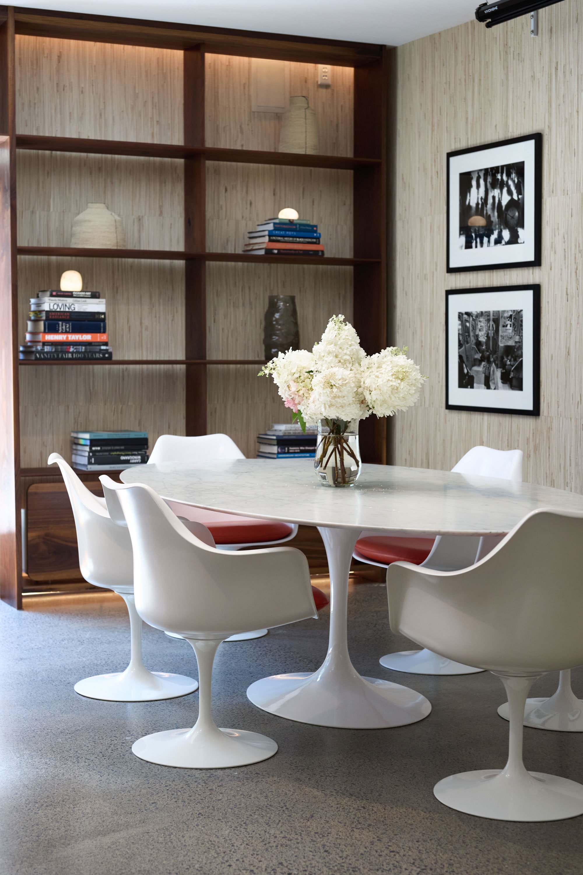 A modern dining room with a white marble table surrounded by white tulip chairs with pink cushions. A glass vase with white flowers is on the table. Behind, a wooden bookshelf holds books and decorative objects. Two black-and-white framed photographs hang on the beige wallpapered wall.