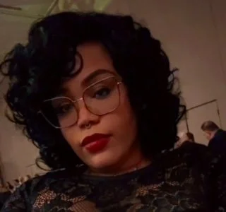 A woman with curly black hair, glasses, and red lipstick taking a selfie indoors.