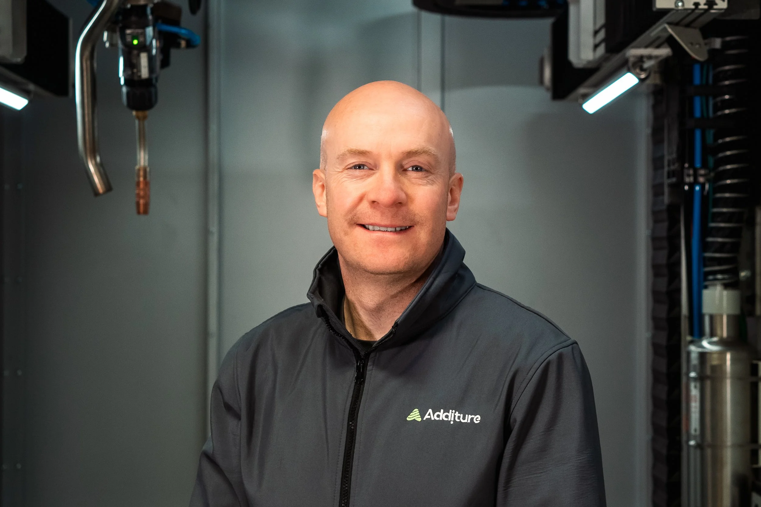 Bald man wearing a gray jacket with an "Additure" logo, standing in a high-tech environment.