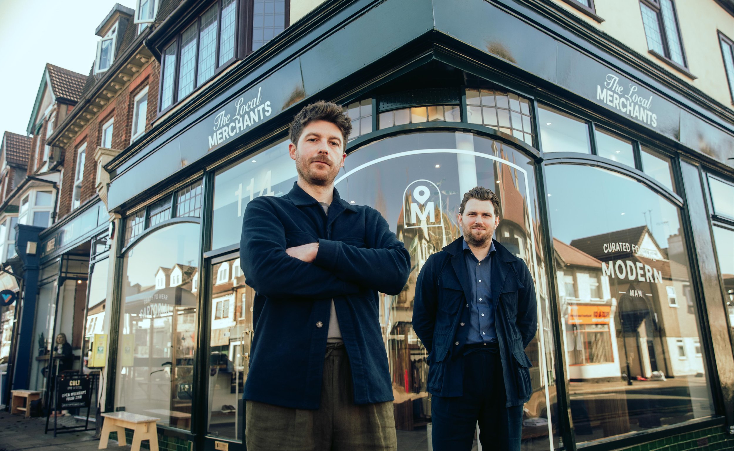 Two men standing in front of "The Local Merchants" storefront on a sunny street.