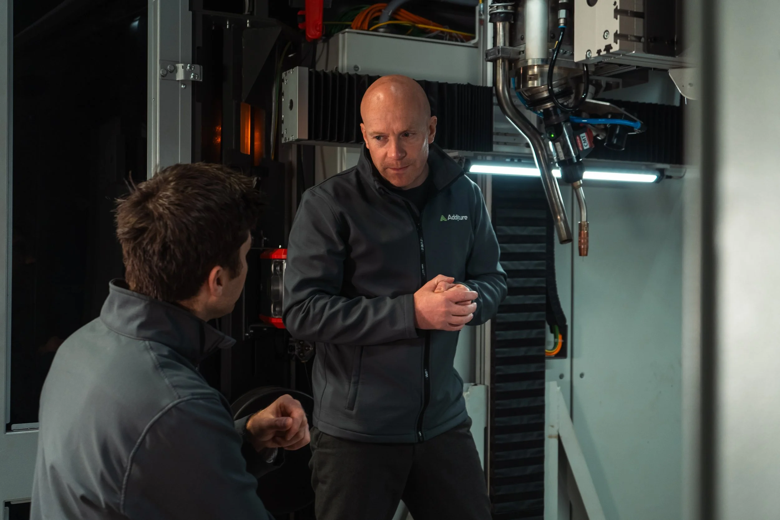 Two men in a technical or industrial setting having a discussion, with machinery in the background. Both are wearing gray jackets with visible branding.