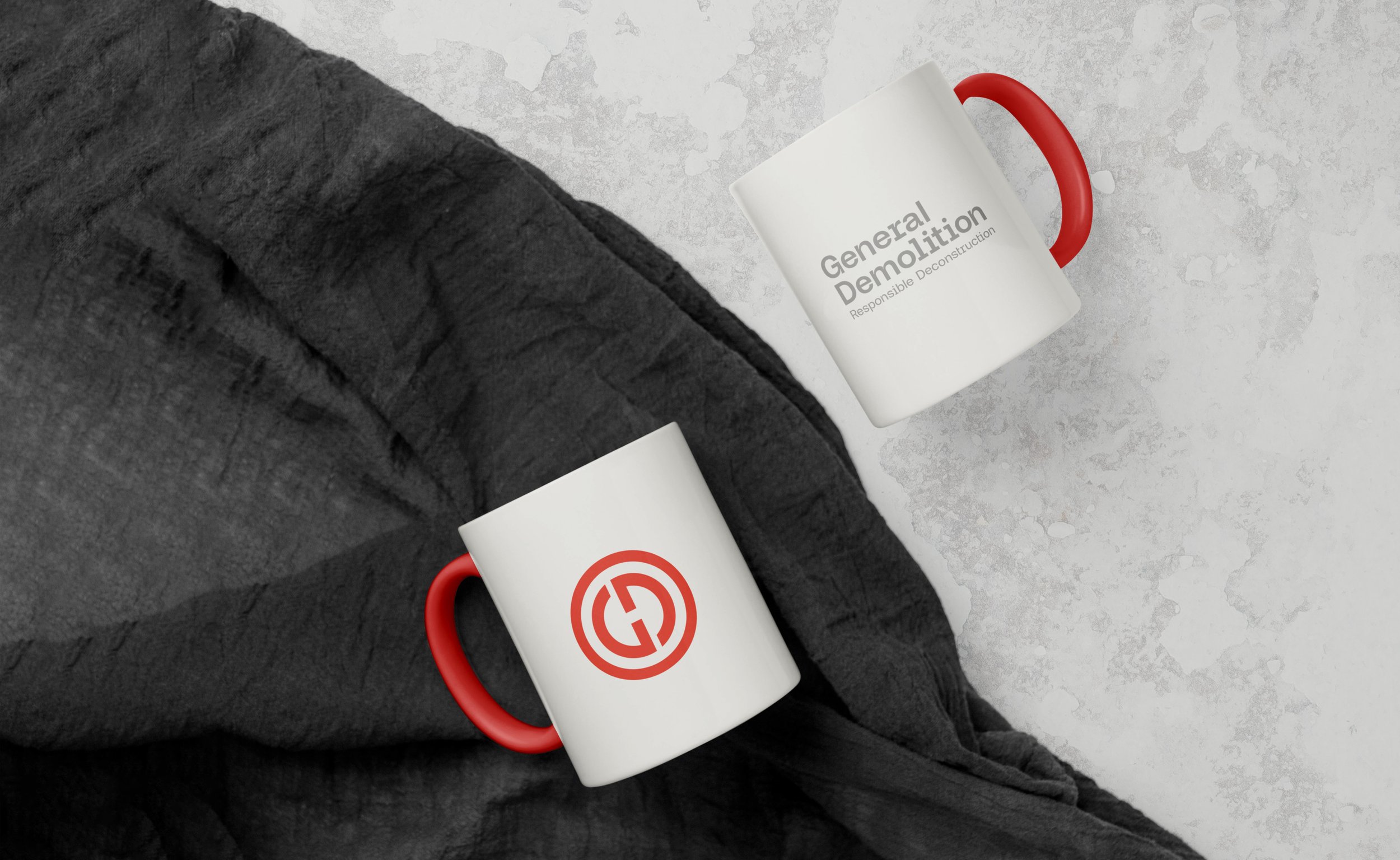 Two white coffee mugs with red handles on a gray textured surface; one mug has "General Demolition" text and the other has a circular logo.
