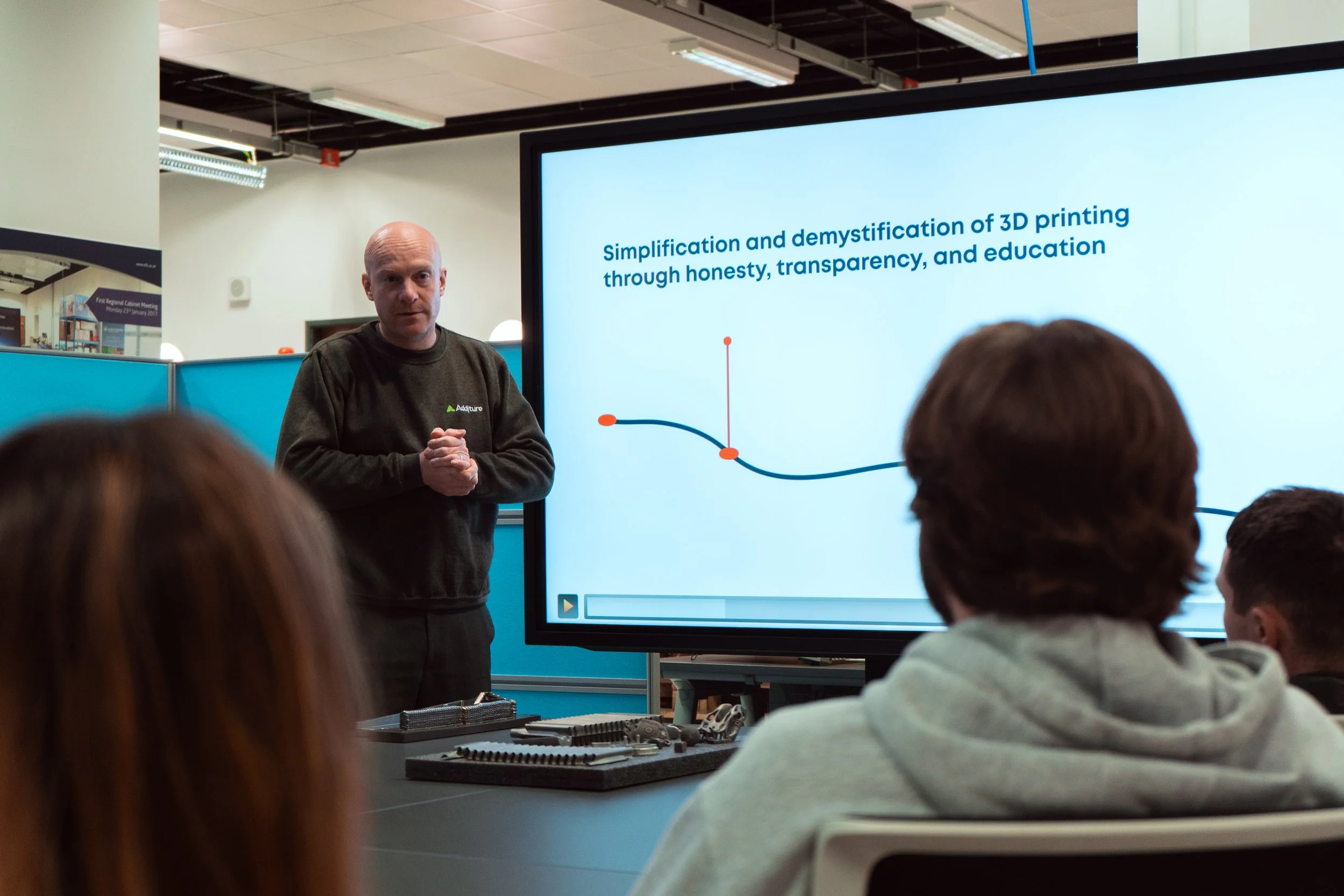 A man presenting to a group with a slide displaying text about 3D printing in an office environment.