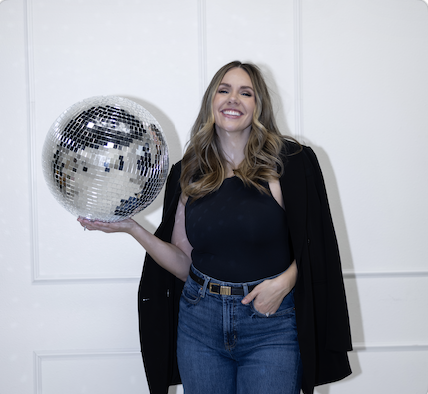 woman smiling holding a disco ball in her right hand, wearing jeans and an open blazer against white wall