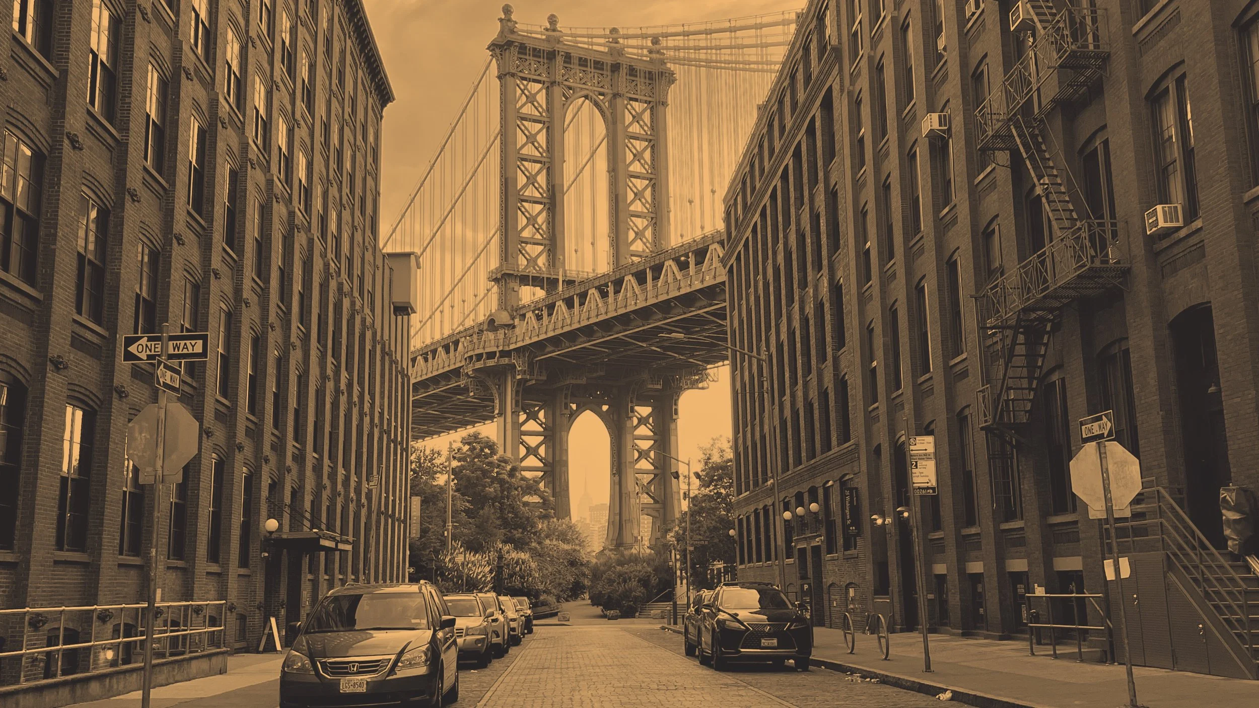 A city street scene with parked cars on both sides, brick buildings with fire escapes, and the Brooklyn Bridge overhead in the background, captured during sunset or sunrise.