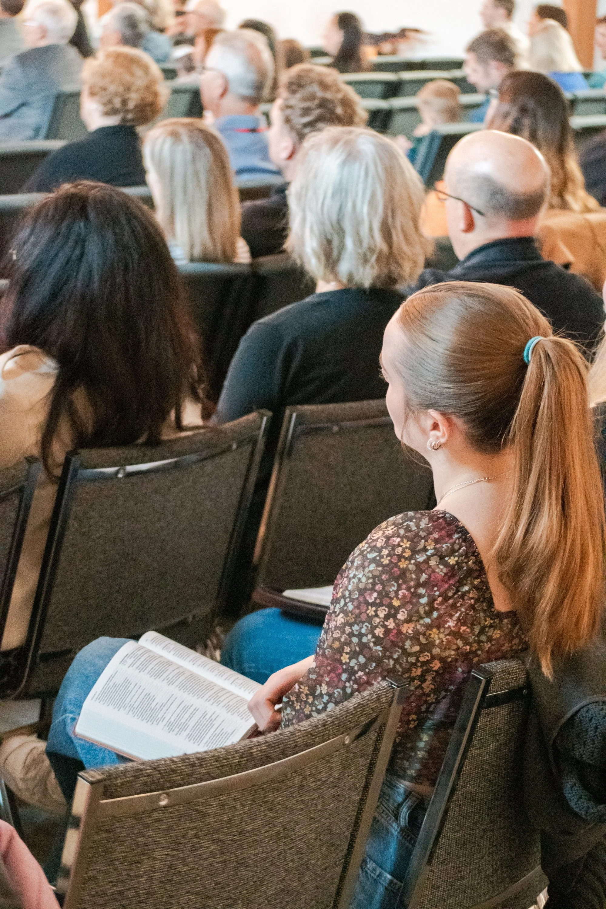 A woman with long hair tied in a ponytail sitting in a church taking notes in a notebook