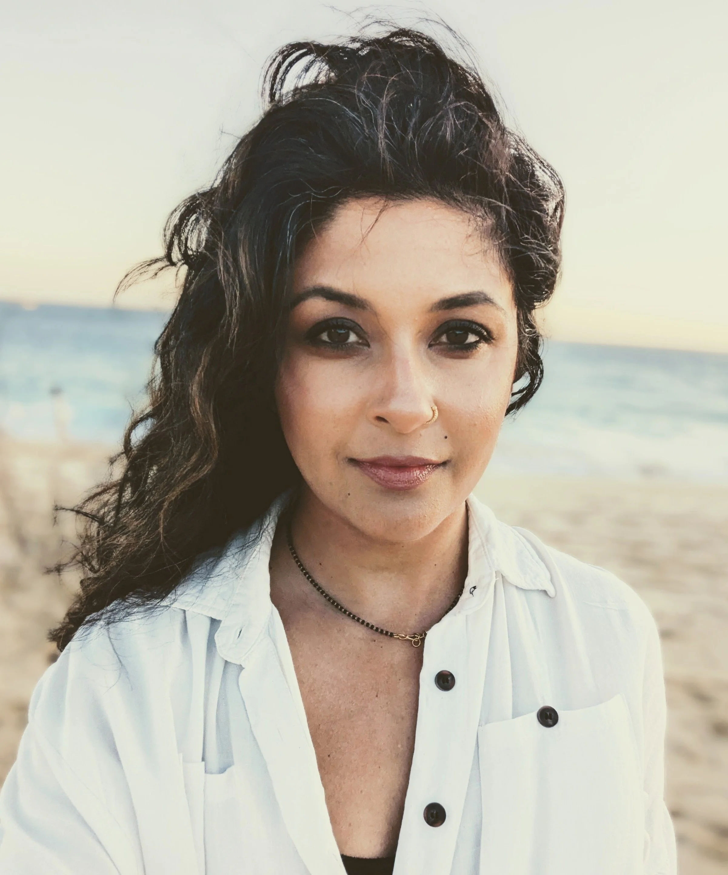 A woman with dark curly hair and a nose piercing, wearing a white shirt, standing on a beach during sunset.