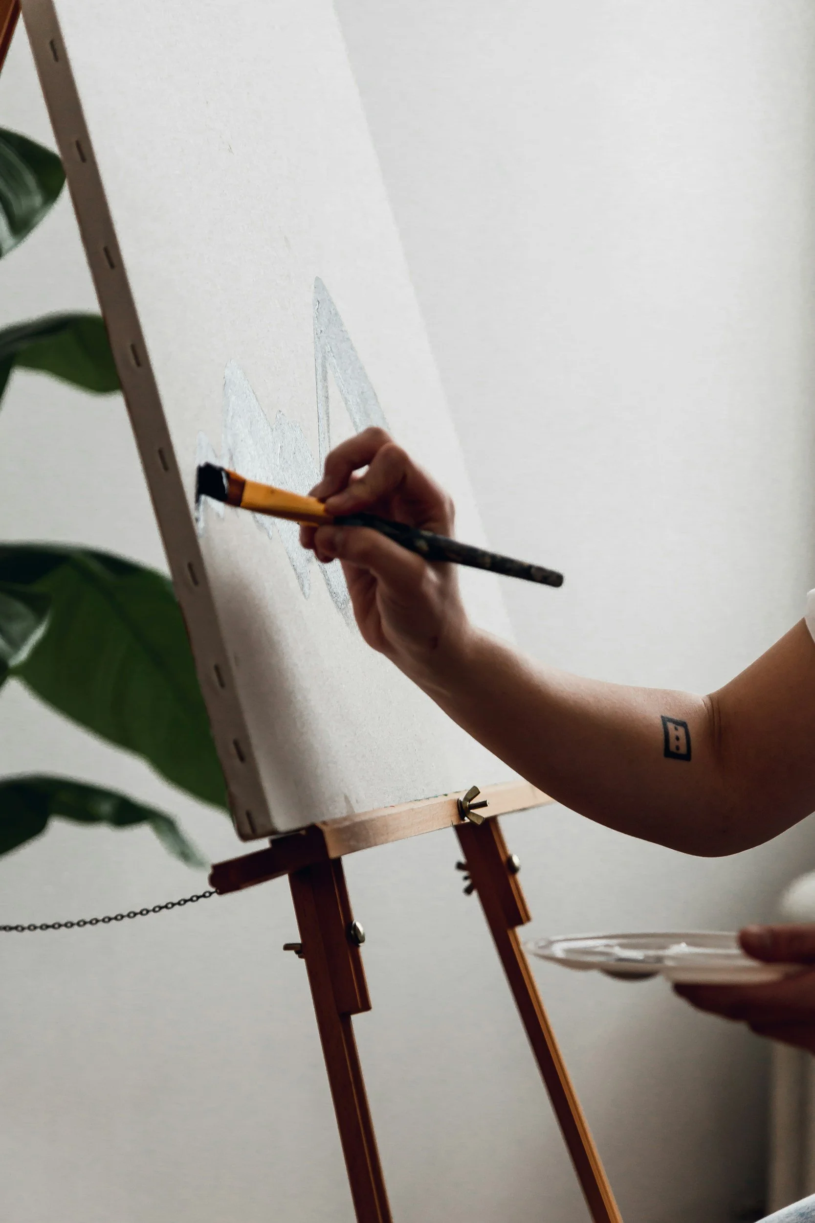 Close-up of a person's hand holding a paintbrush, painting mountain shapes on a white canvas or paper on an easel.