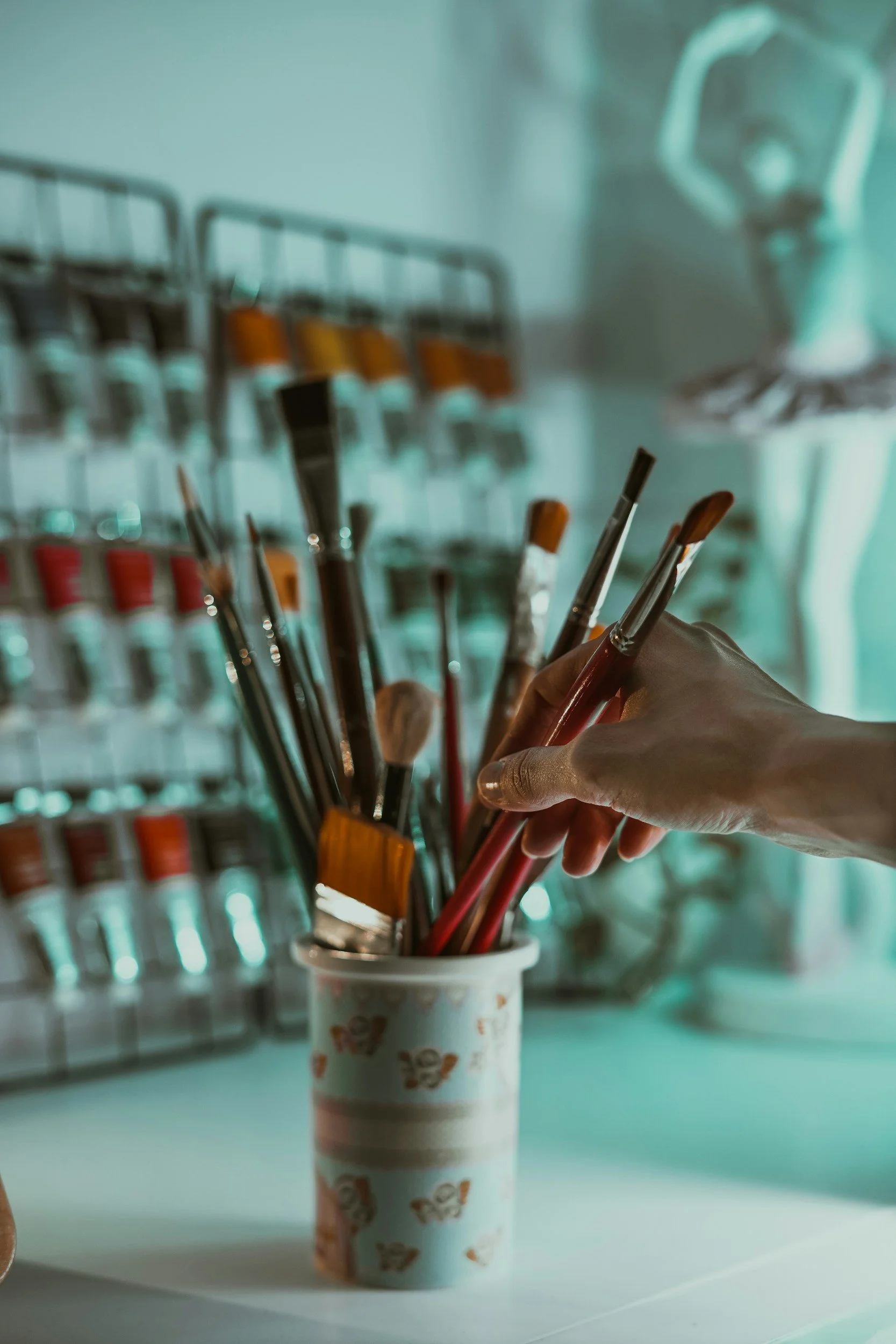 A hand holding paintbrushes inside a paper cup with ice cream mascot prints, with a blurred background of wall-mounted brushes and a spray gun.