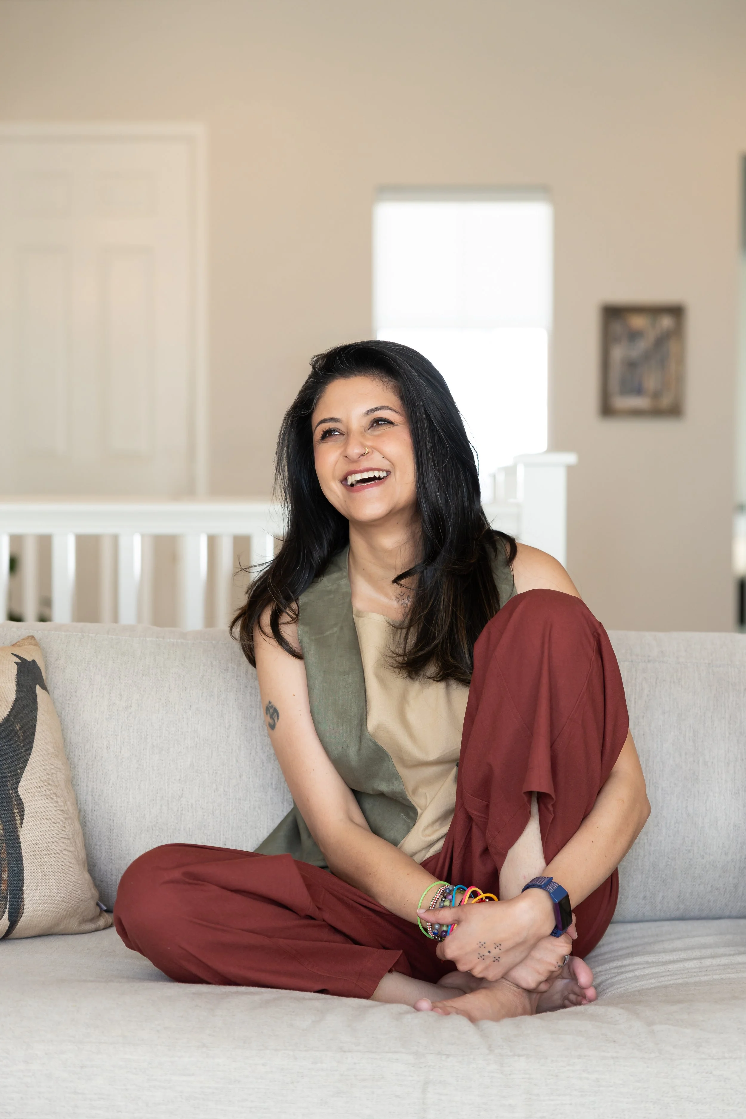 A woman with long dark hair, wearing a sleeveless beige and green top with maroon pants, is sitting cross-legged on a beige couch, smiling and looking off to the side. She's accessorized with colorful bracelets and a nose ring. The background shows a beige wall, a window with sunlight, and a framed picture.