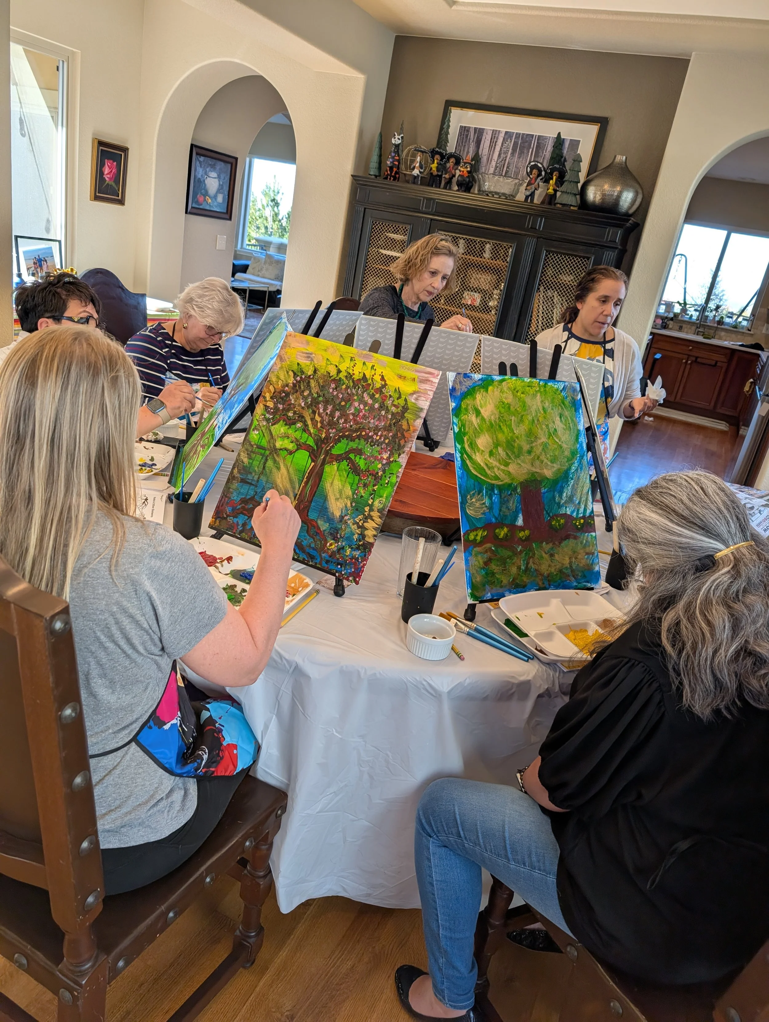 People painting landscape pictures on canvases during a group art class in a cozy home setting.