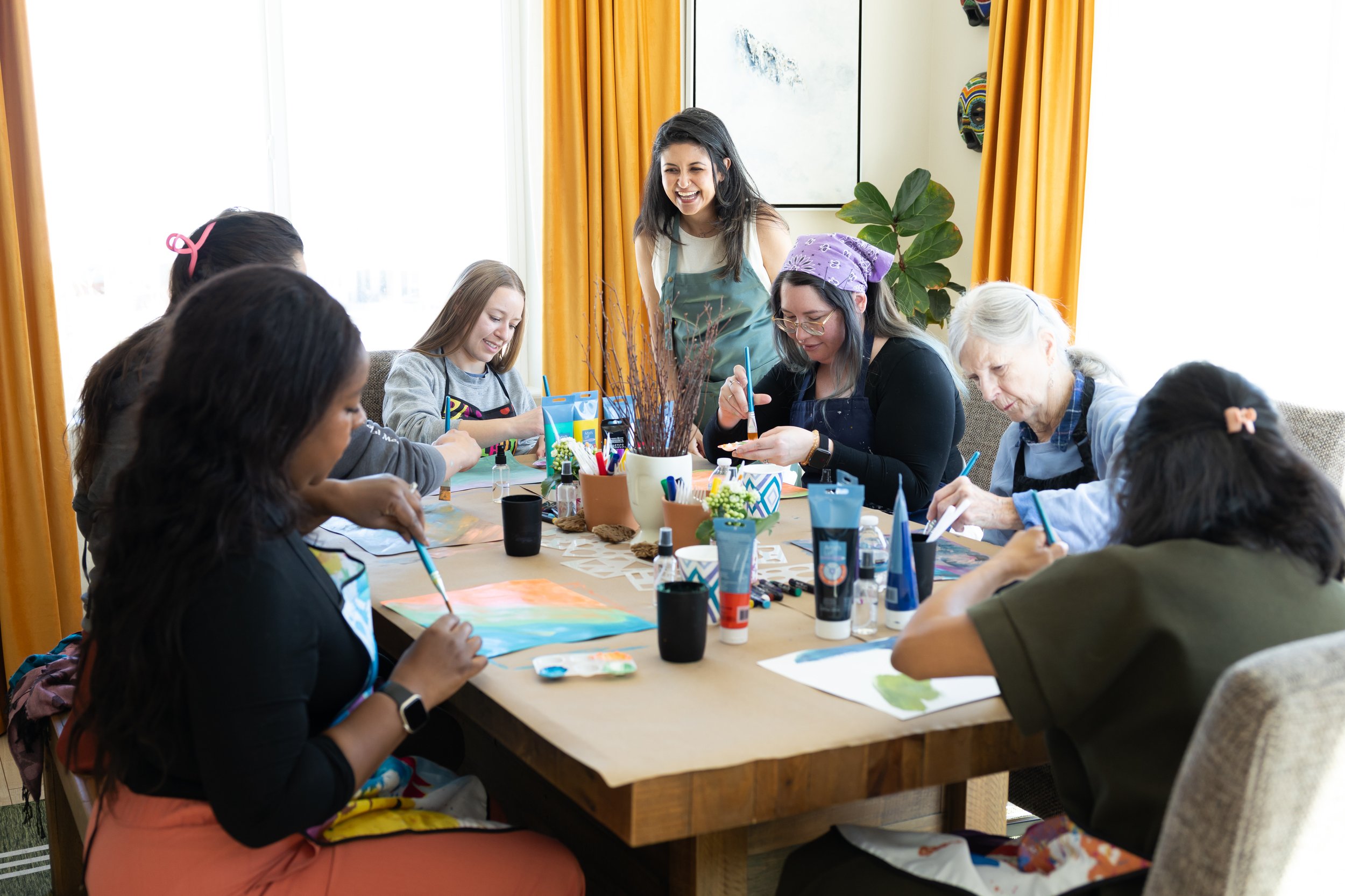 A group of women and girls sitting around a table engaged in painting or craft activity. A woman in the center is smiling and wearing a lavender headscarf and glasses. The room has large windows with yellow curtains, a large plant, and artwork on the wall.