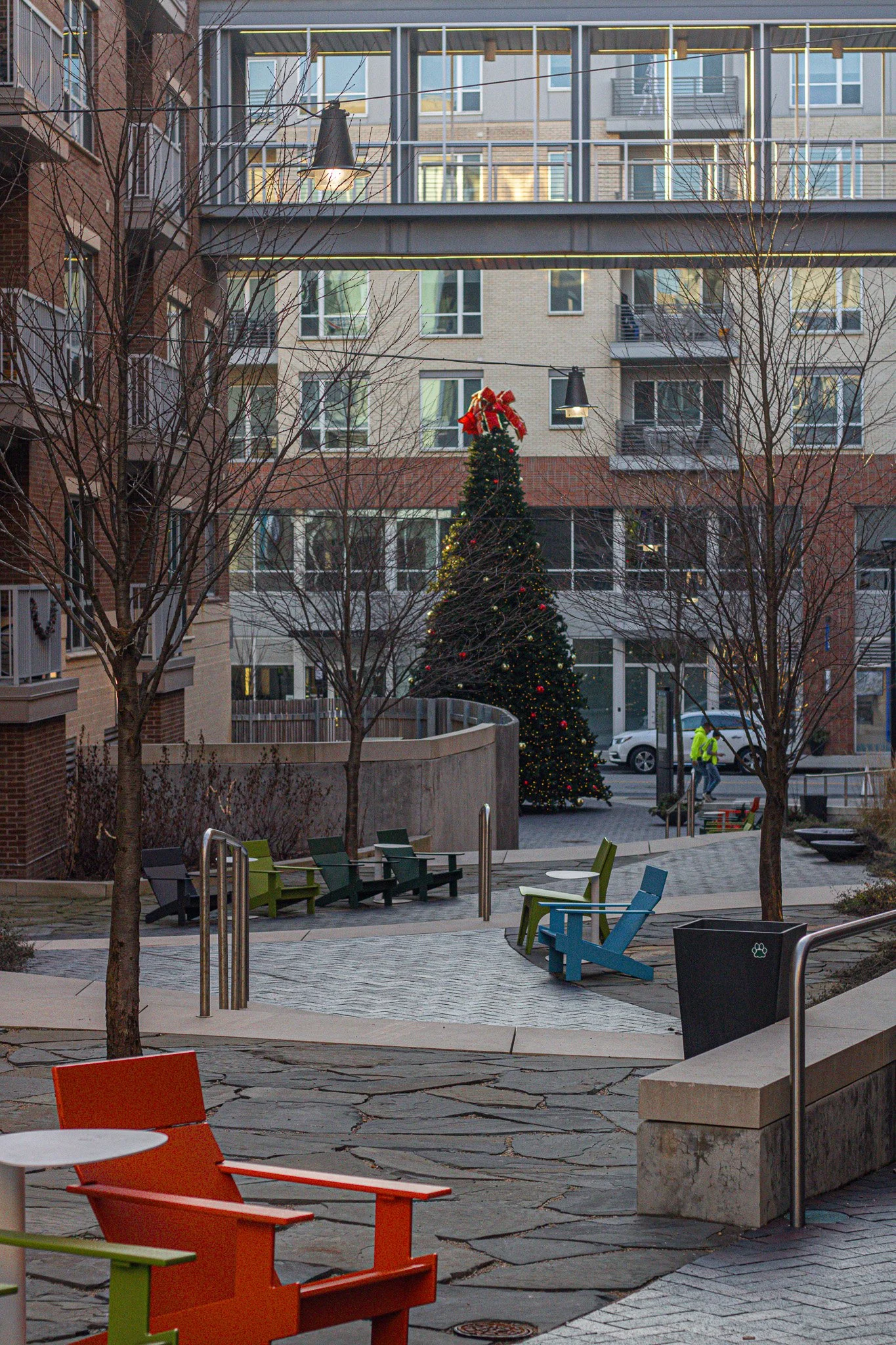 An outdoor urban courtyard with colorful chairs, leafless trees, and a decorated Christmas tree with a red bow on top, surrounded by apartment buildings.