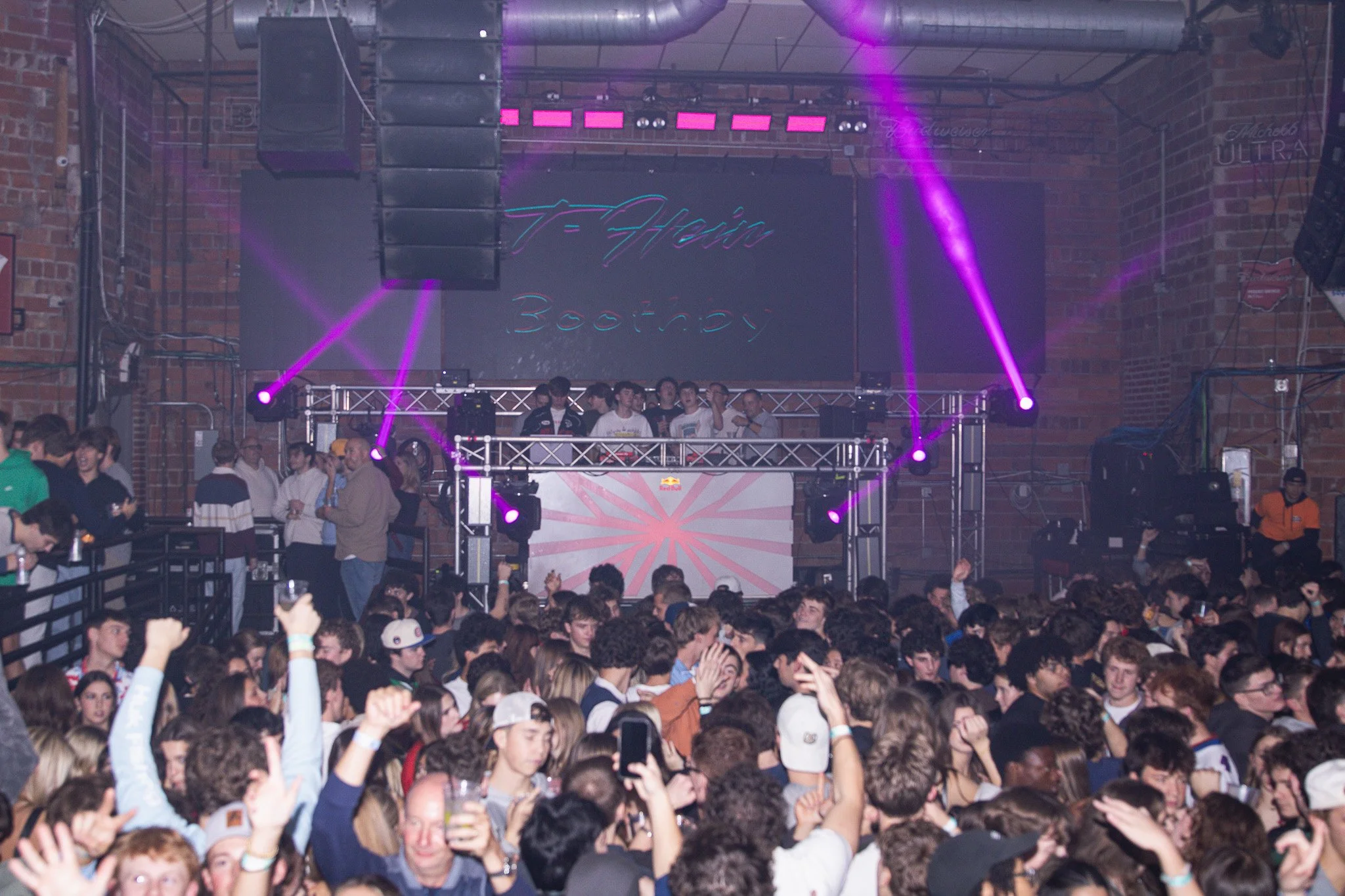 Crowd of young people at a DJ concert in a nightclub, with people dancing and taking photos, a DJ on stage with lighting and large screen in the background.