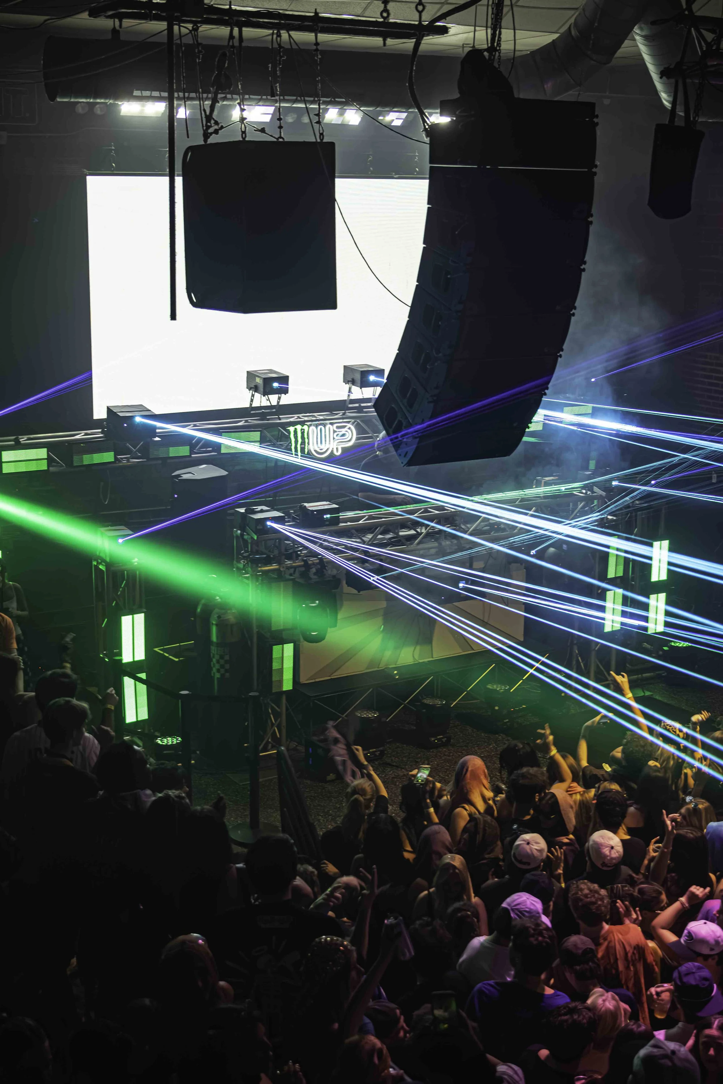 Crowd at a nightclub dance floor with green laser lights, large speaker system, and a DJ booth with MONSTER energy drink logo in the background.