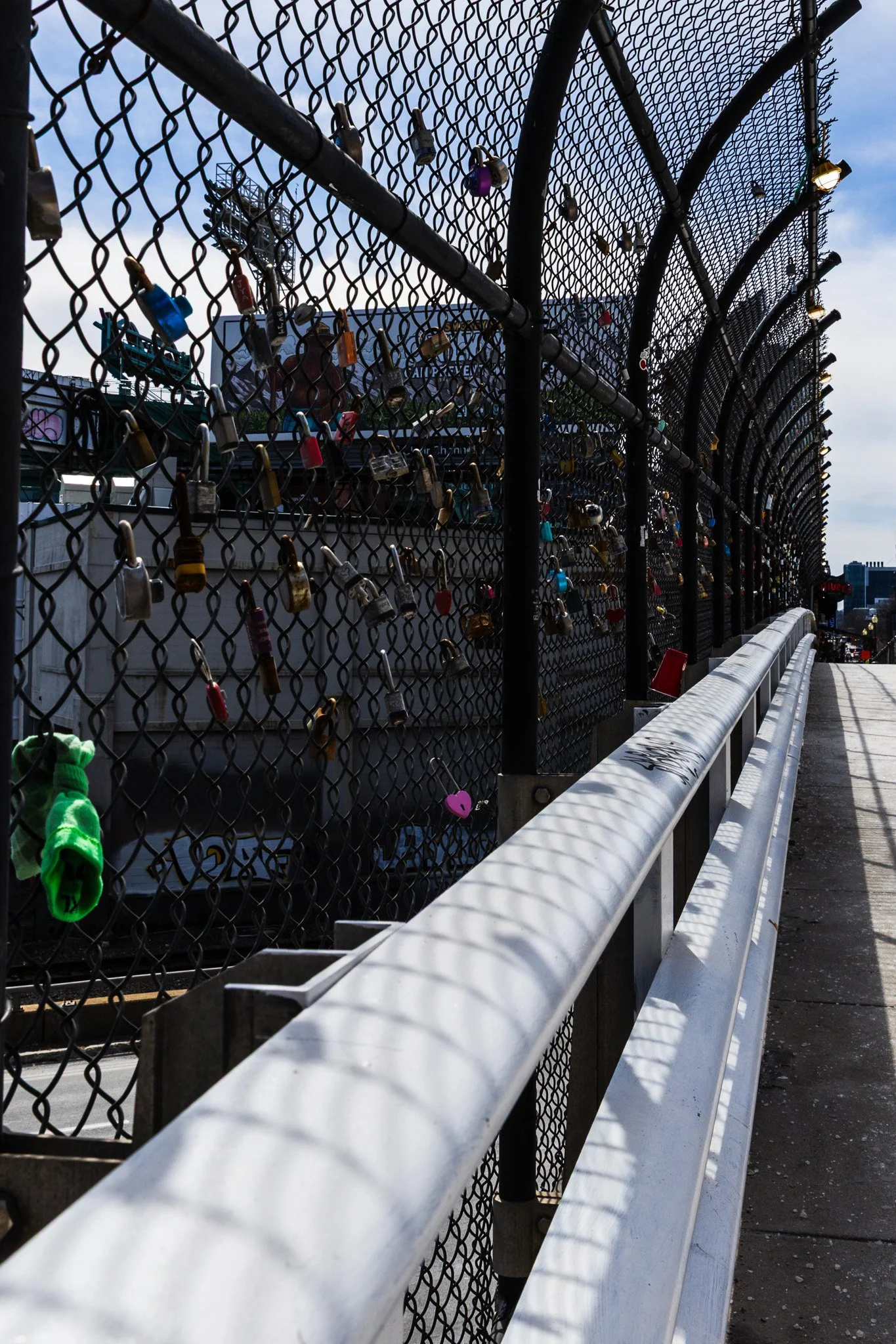 City street view with a fence covered in numerous locks, possibly love locks, along a bridge or walkway, with city buildings in the background and a partly cloudy sky.