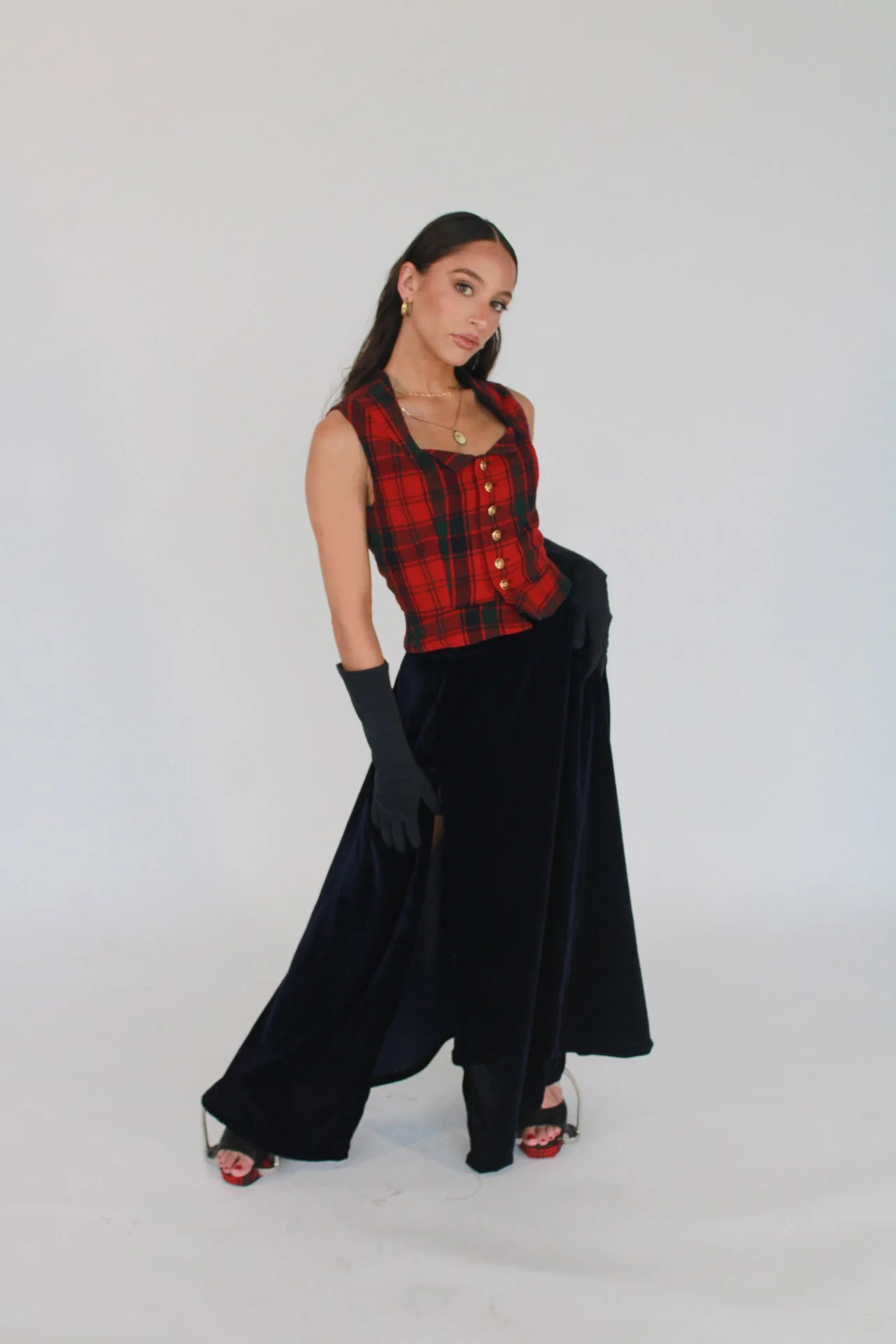 Woman in black gloves, red plaid sleeveless top, and wide-leg black pants standing against a white background.